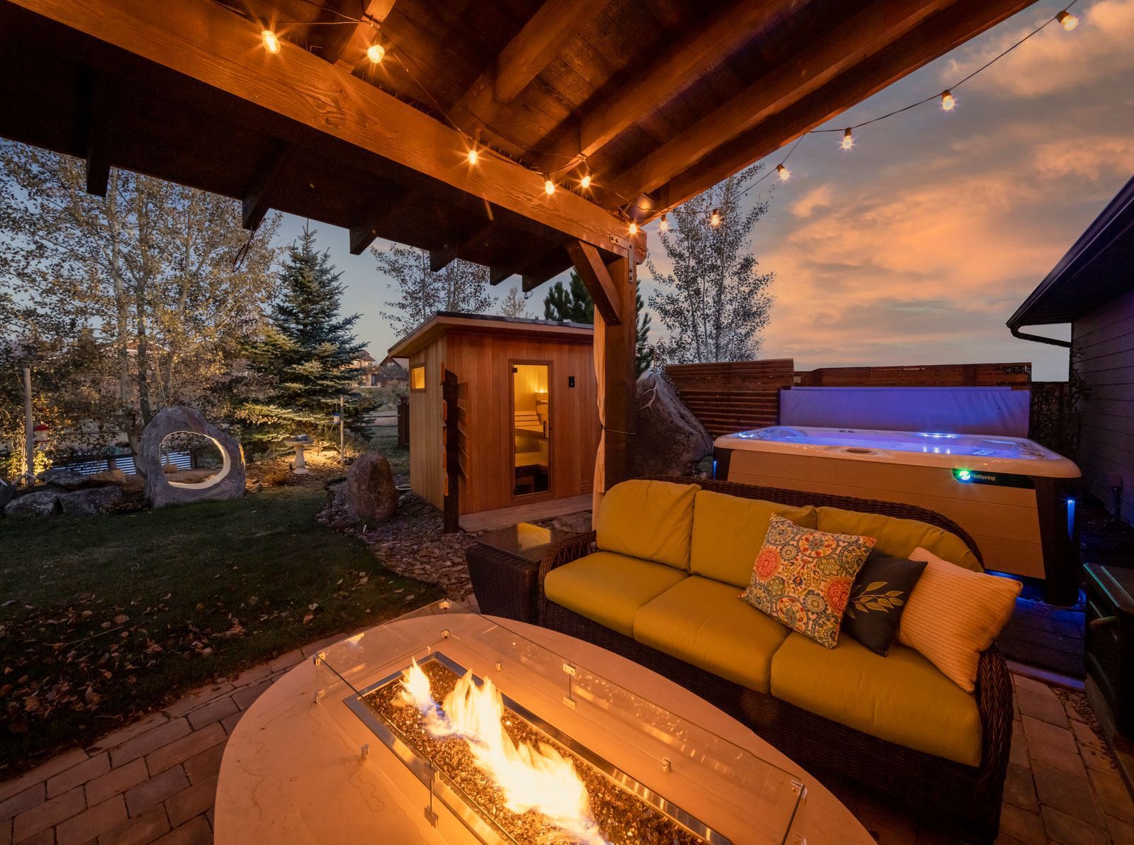 Cozy patio with lit fire pit, sofa, hot tub, and sauna under a wooden pergola at dusk.