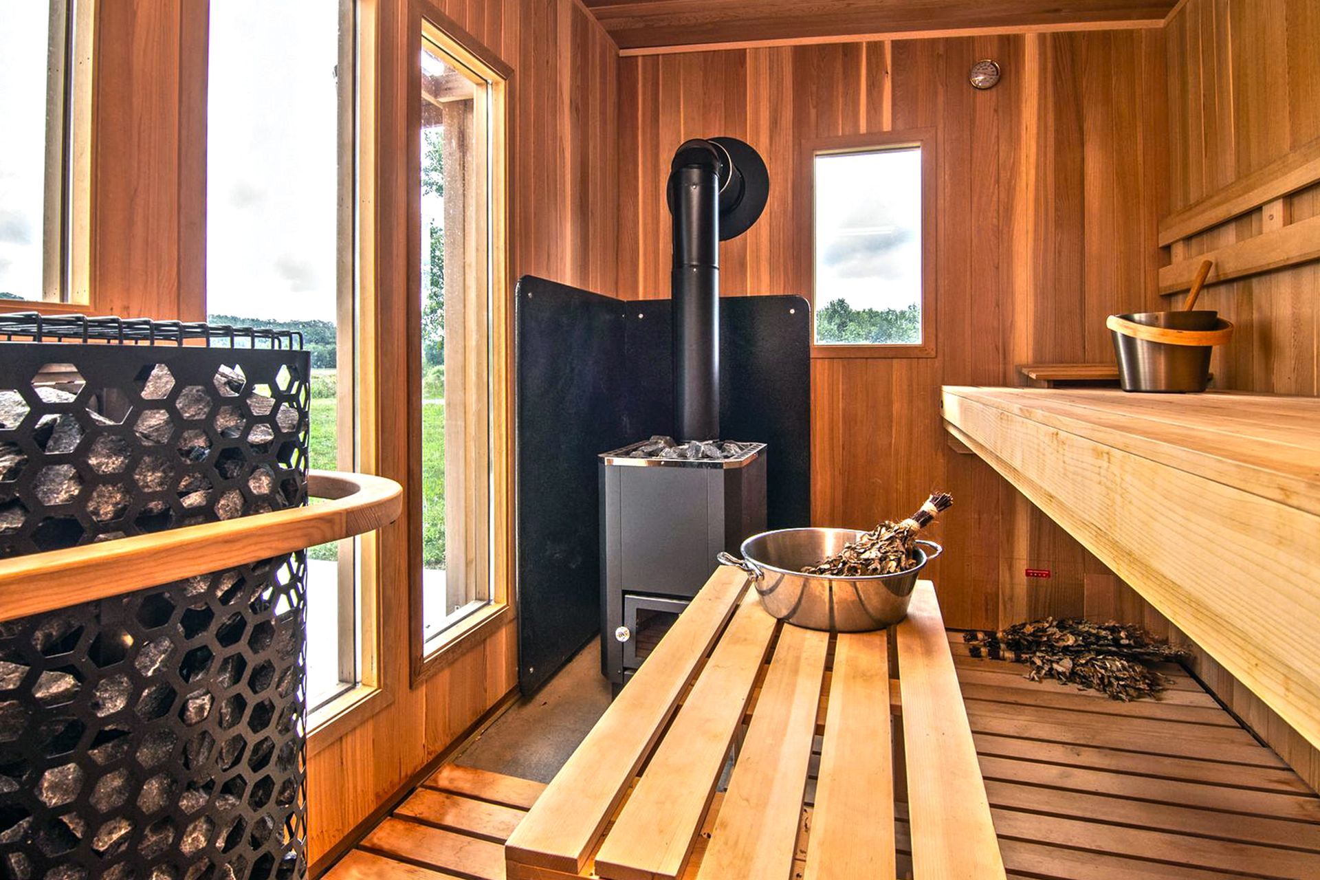 Wooden sauna with a stone heater, benches, and windows with a view of the outdoors.