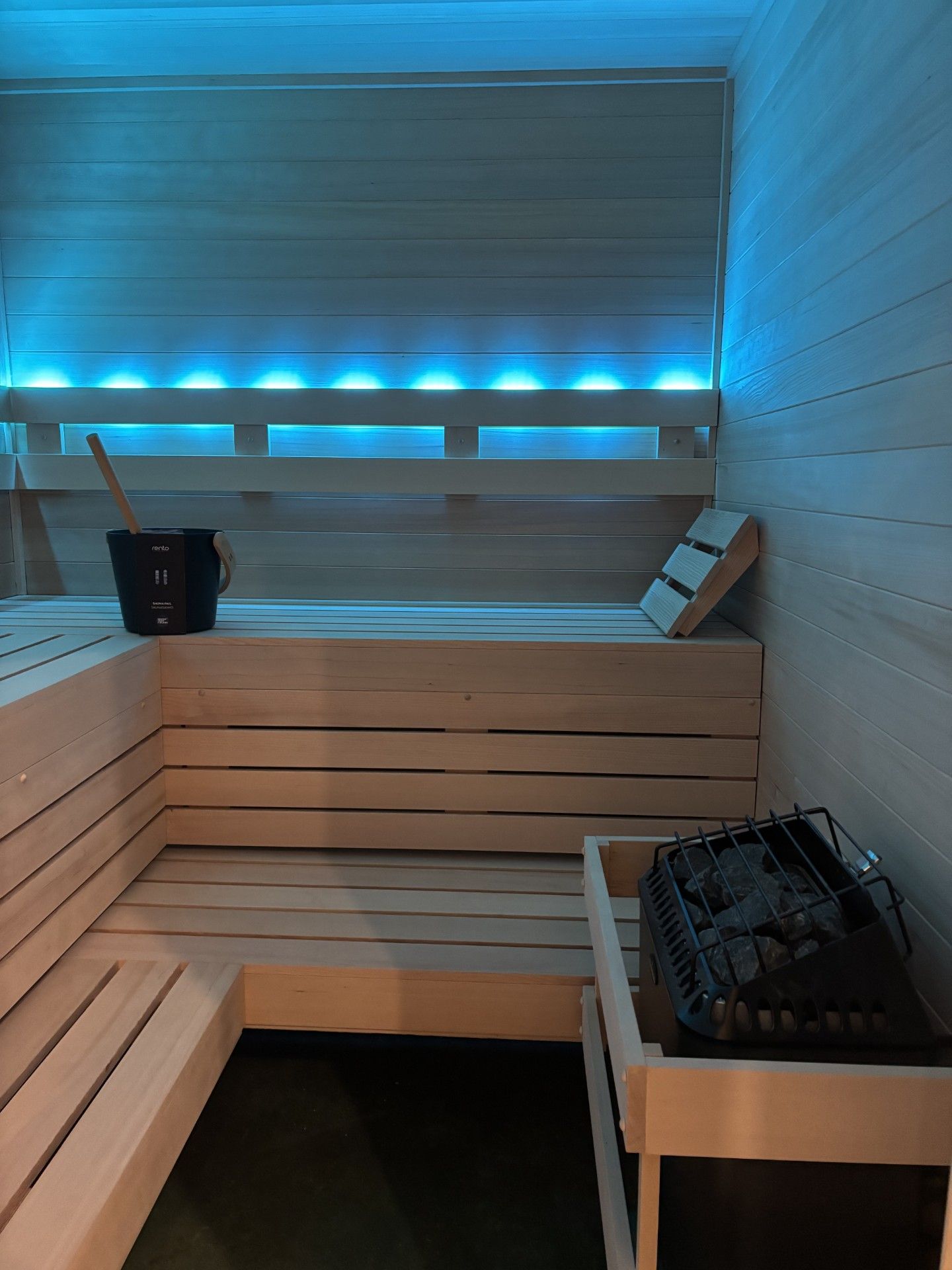 Inside a light-wood sauna with blue LED lighting; bucket and ladle, heater, and benches.