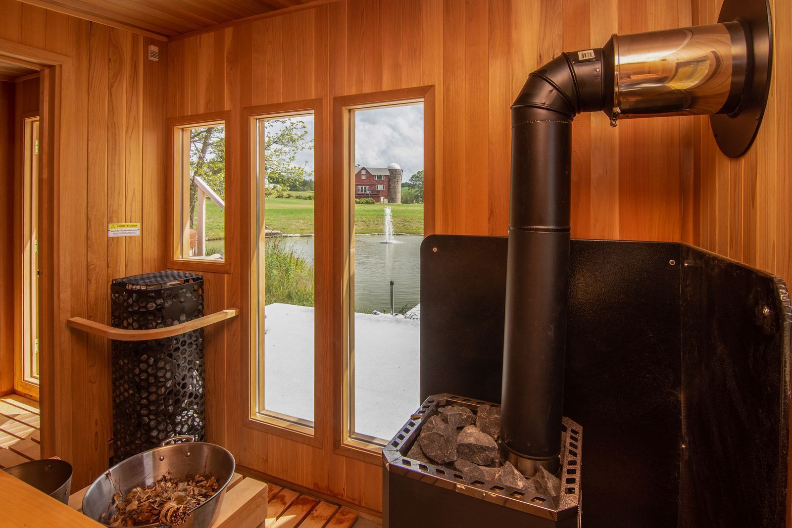 Wood-paneled sauna with windows overlooking a snowy landscape. A stove with a chimney, a bucket of rocks, and a wooden bucket are inside.
