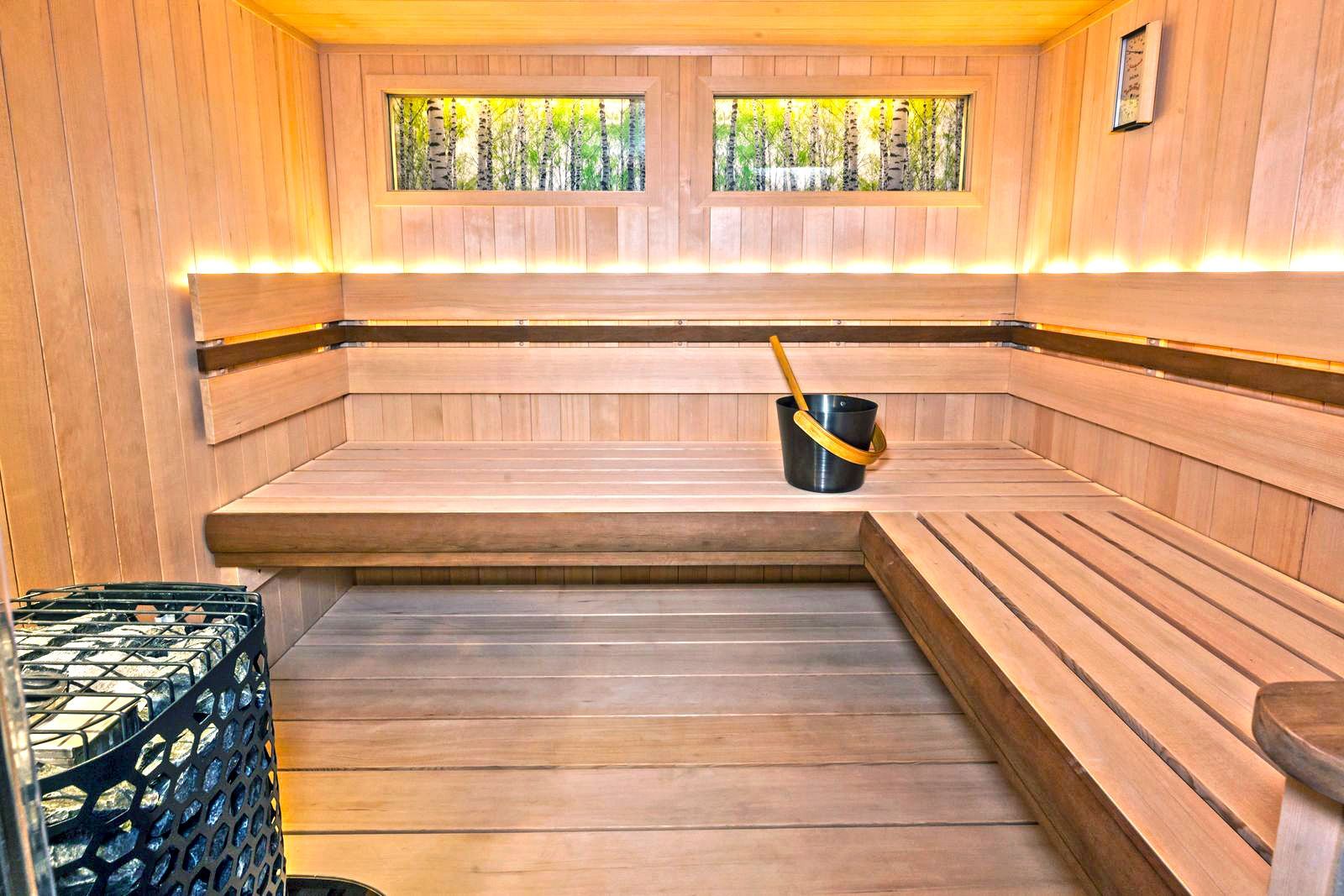 Wooden sauna with benches, bucket, and a heater. Sunlight streams in from window.
