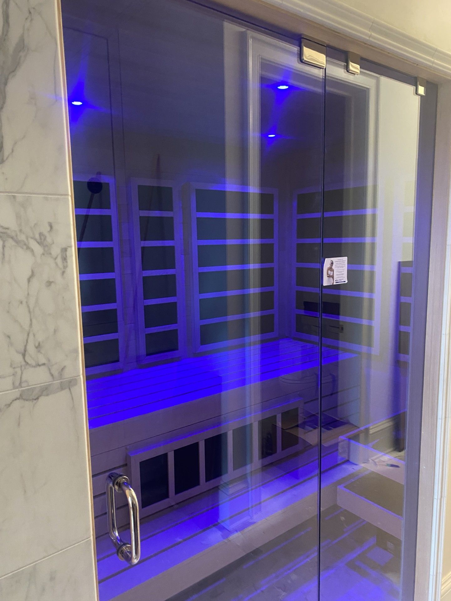 Glass-doored sauna with blue interior lighting. Wooden benches, rectangular panels, and marble wall.