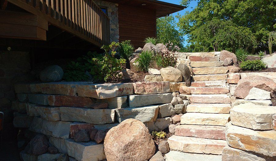 Stone retaining wall with steps leading up to a wooden deck, plants and trees in the background.