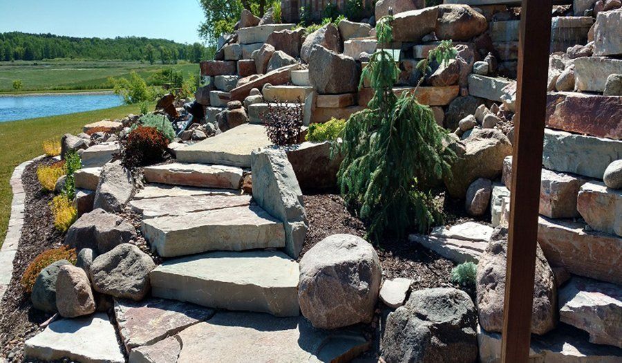 Stone steps and rock garden by a lake, lush greenery, and sunny setting.