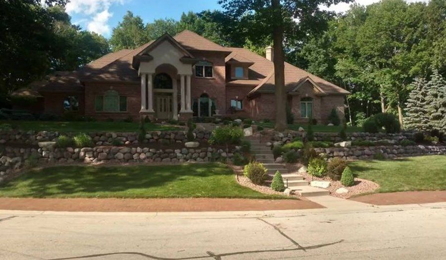 Brick house with pillars, brown roof, and landscaped yard. Steps lead to the front door; trees in background.