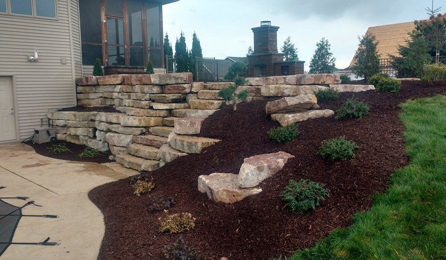 Stone steps lead up a sloped landscape with mulch, shrubs, and a house in the background.