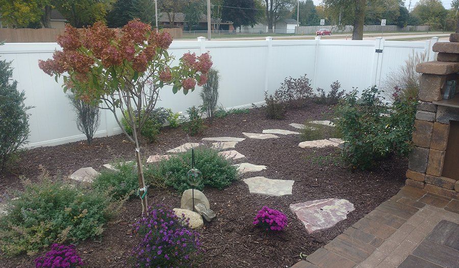 Landscaped front yard with stepping stones, shrubs, flowers, and a tree, against a white fence.