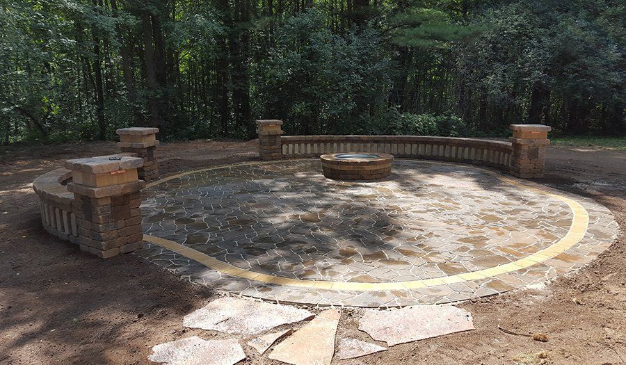 Circular stone fire pit area with seating and posts in a wooded setting.