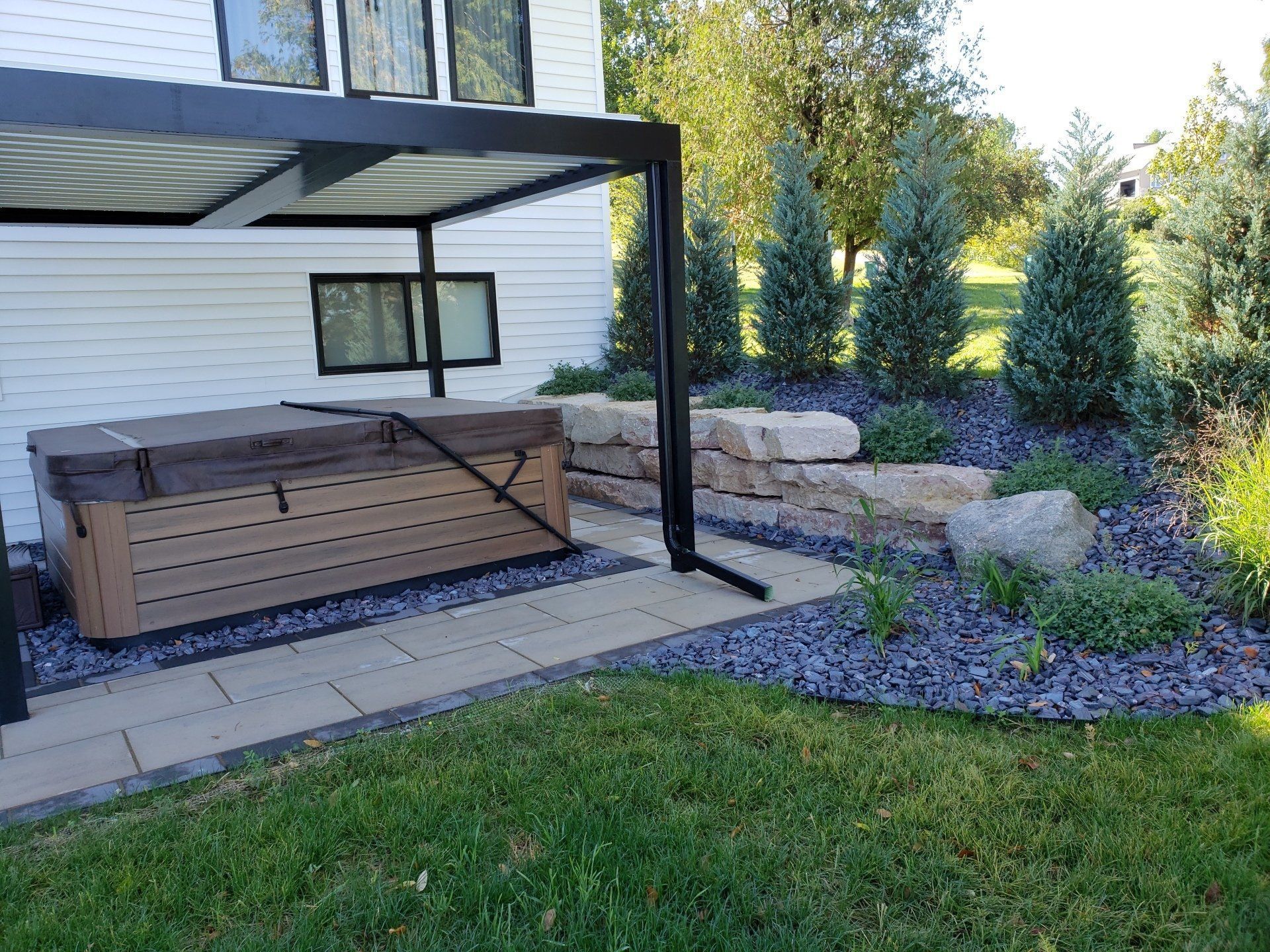 Hot tub with cover under a pergola, beside a stone wall and landscaping with trees, gravel, and grass.