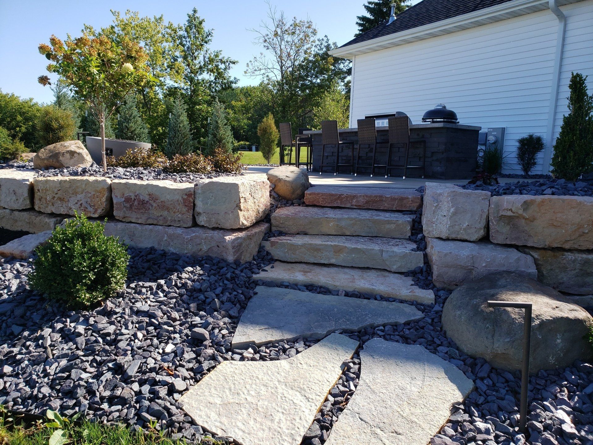 Stone steps leading up to a patio with outdoor furniture, surrounded by landscaping and a stone retaining wall.