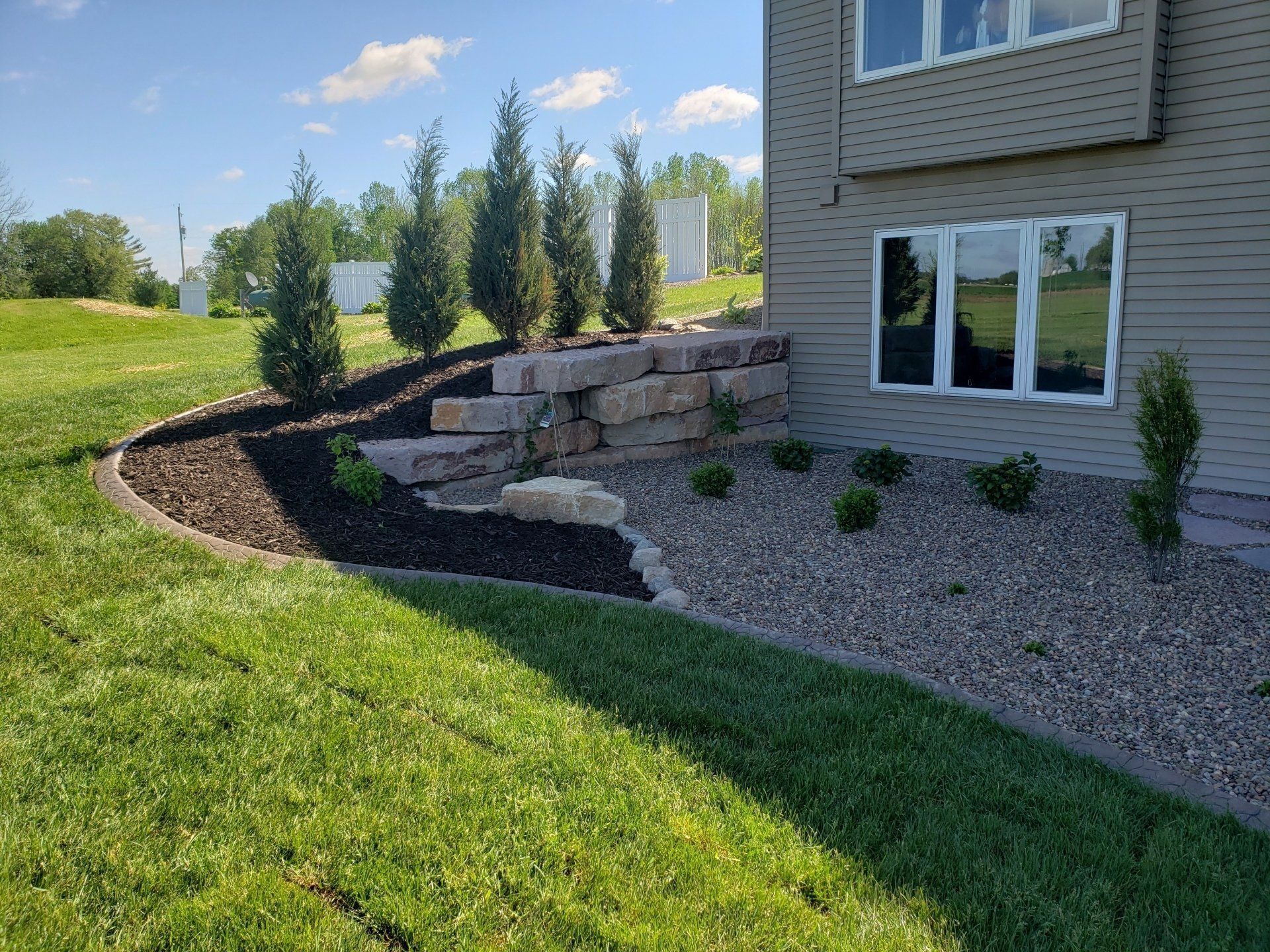 Landscaped yard with retaining wall, trees, and dark mulch beds next to a beige house, green grass in foreground.