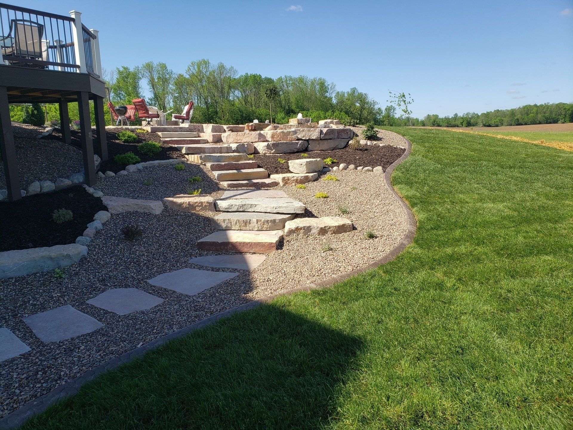Stone steps and landscaping leading up a hillside, with a deck on the left and a green lawn on the right.