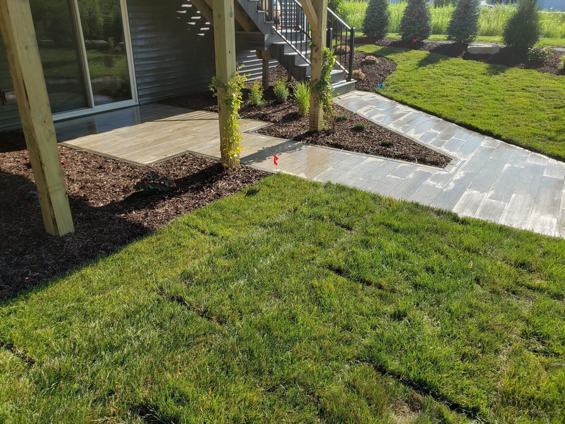 Lush green lawn and stone walkway lead to a house with wooden beams, stairs, and mulch beds.