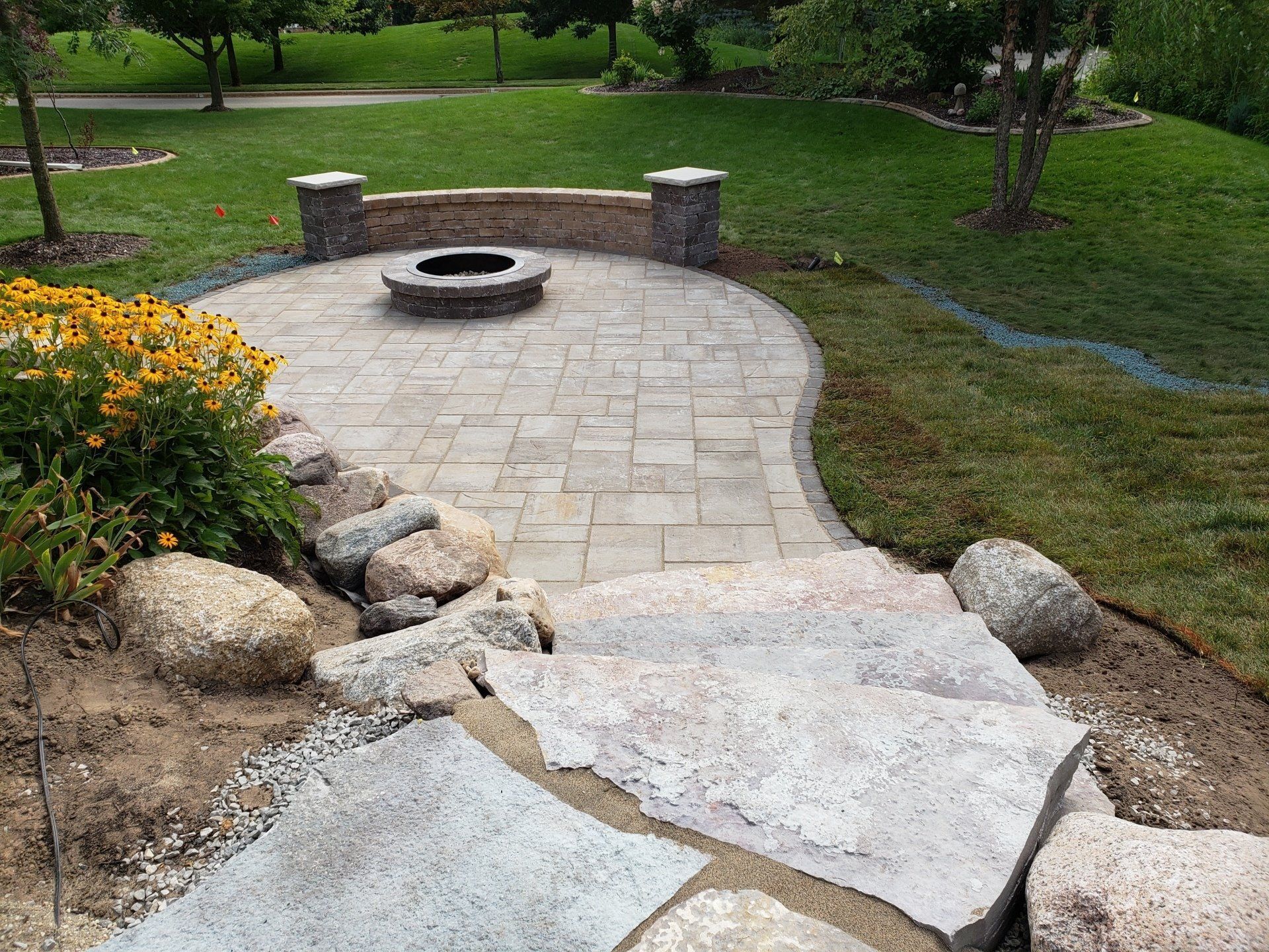 Stone patio with fire pit and built-in seating, accessed by stone steps, in a grassy backyard.
