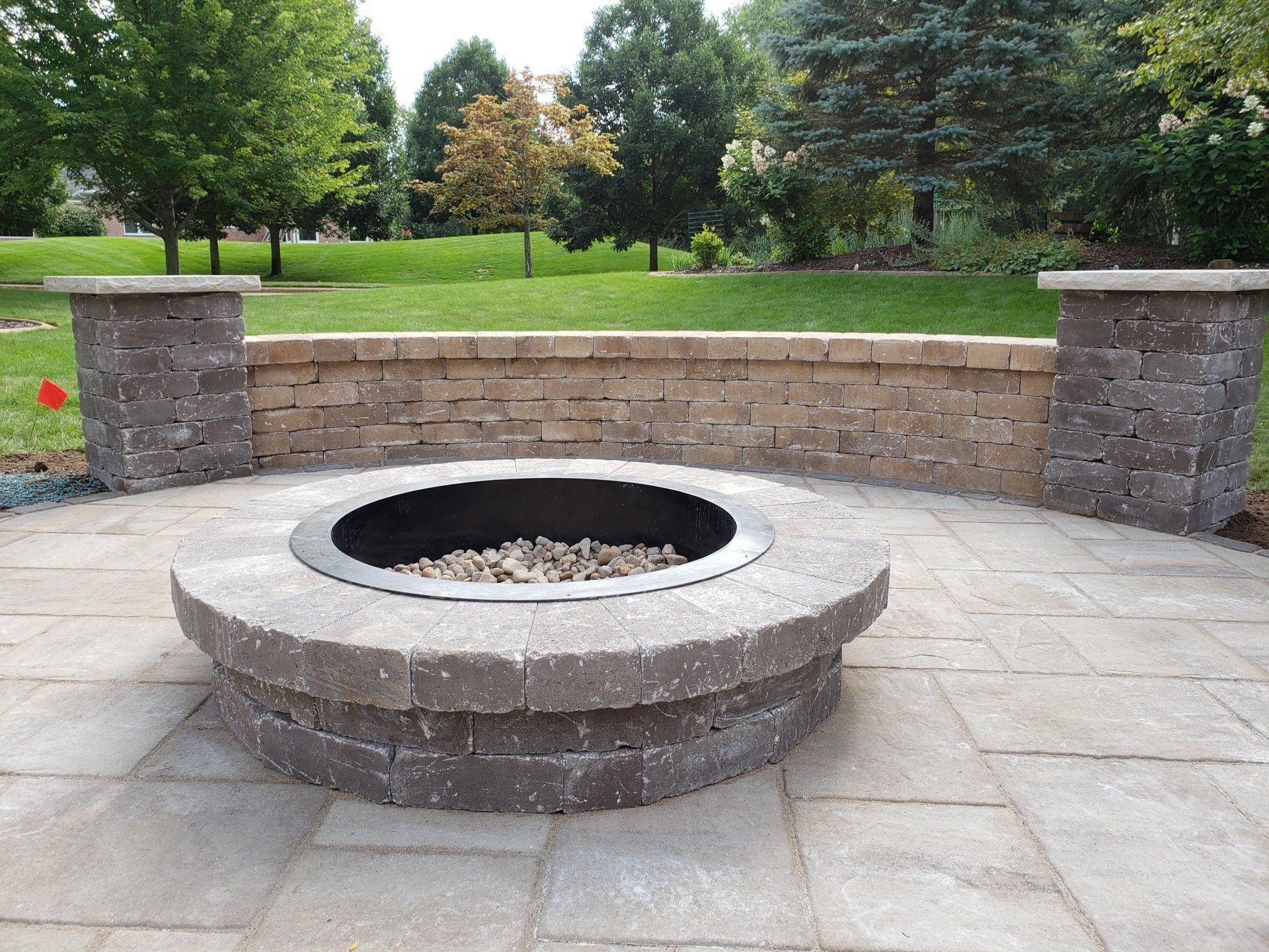 Fire pit on a stone patio with a low retaining wall and pillars in a yard.
