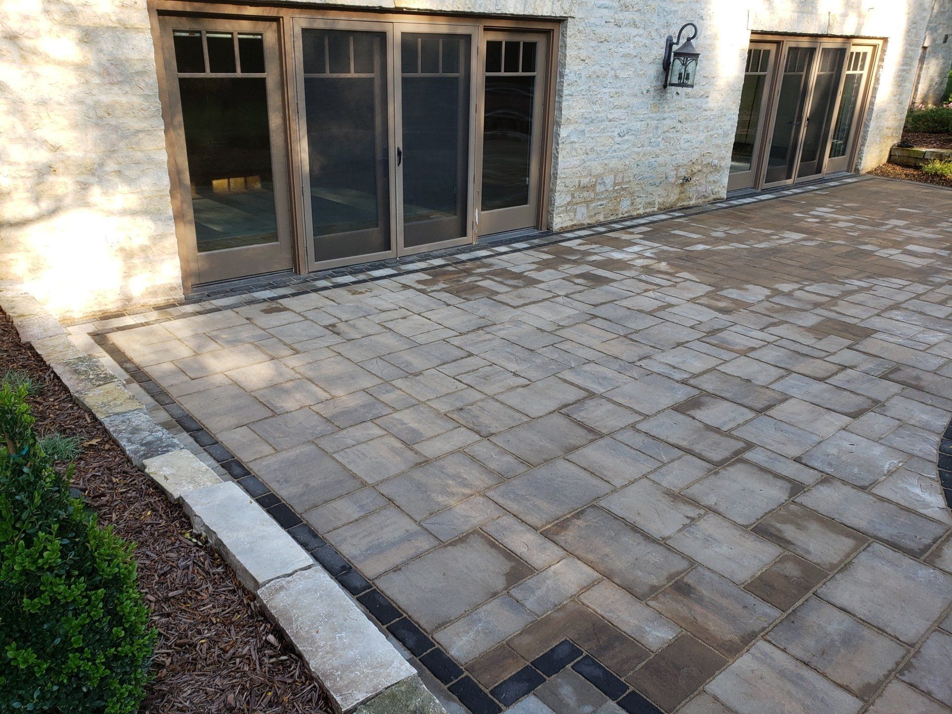 Stone patio outside a building with brown framed glass doors. Brick border and landscaping.