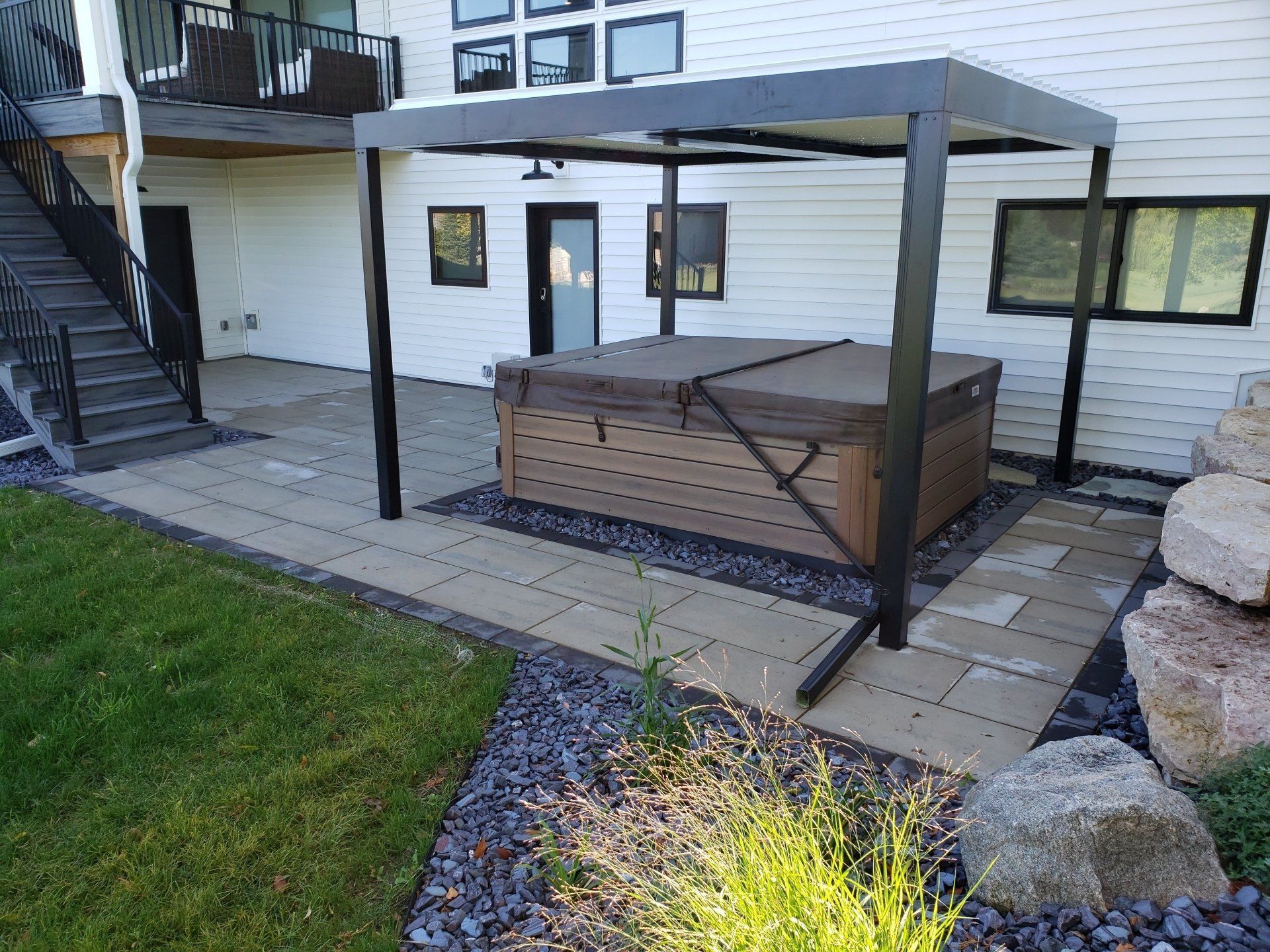 Hot tub under a black pergola on a stone patio next to a house with a staircase and landscaping.