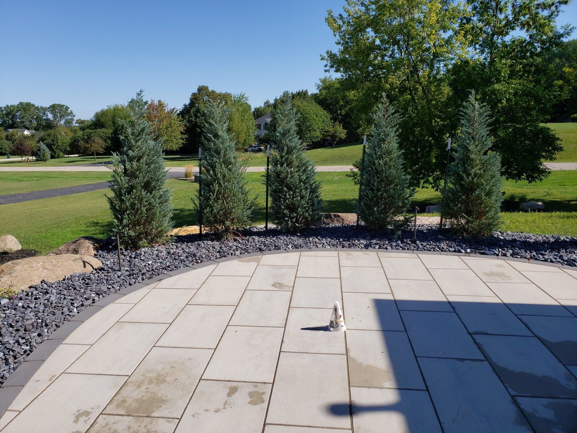 Stone patio with line of evergreen trees and black rocks. Green lawn in background.