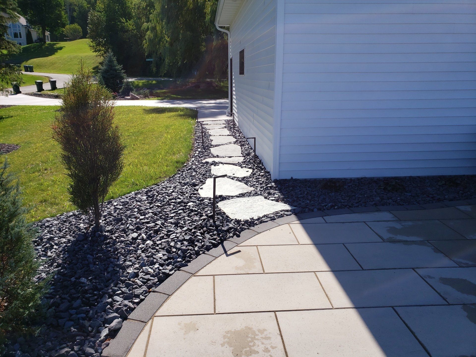 Stone pathway next to a white building. Black rock borders the pathway with green grass and trees.