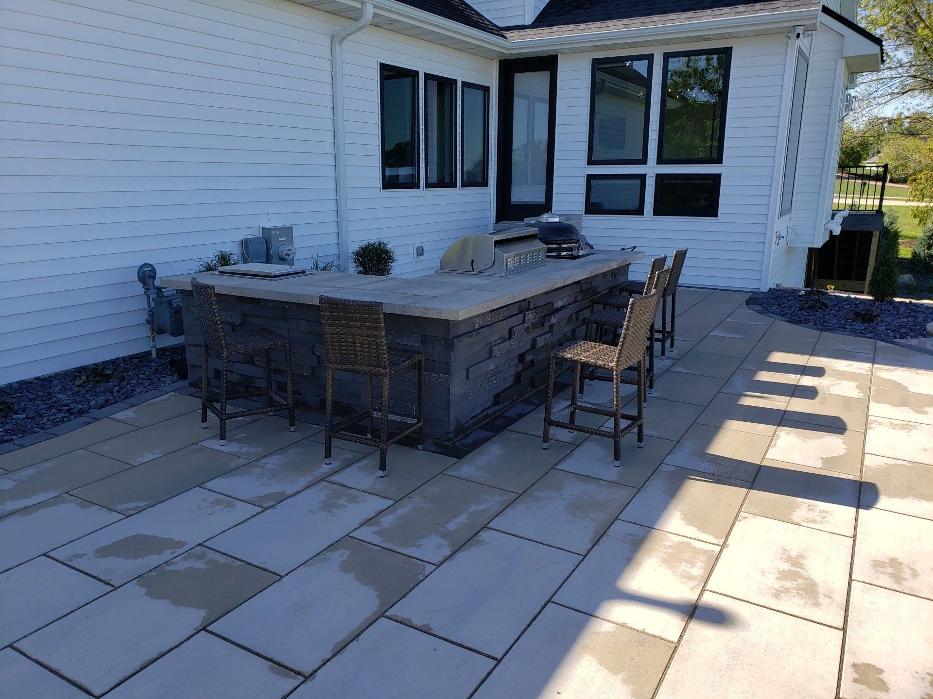 Outdoor kitchen with a bar, seating, and grill on a stone patio next to a house.