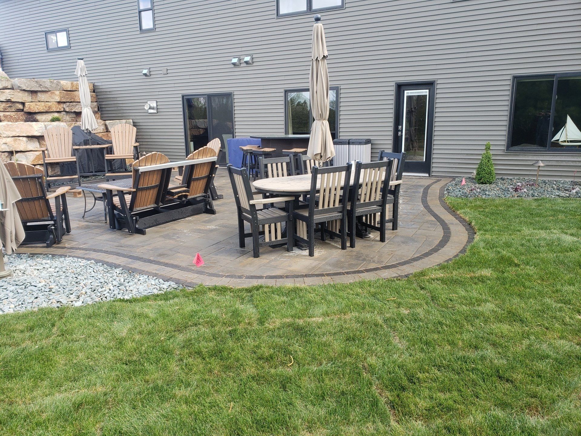 Patio with seating, umbrella, and table. Grass and decorative stone border. House exterior in background.