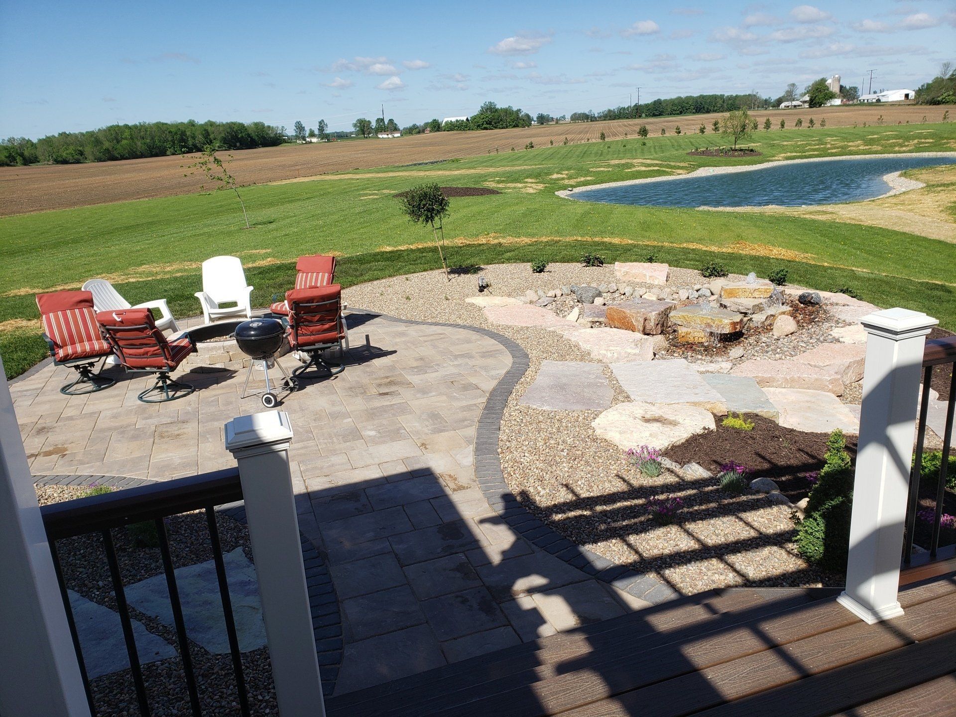 Patio with seating, grill, and stone landscaping overlooking a pond and fields under a sunny sky.