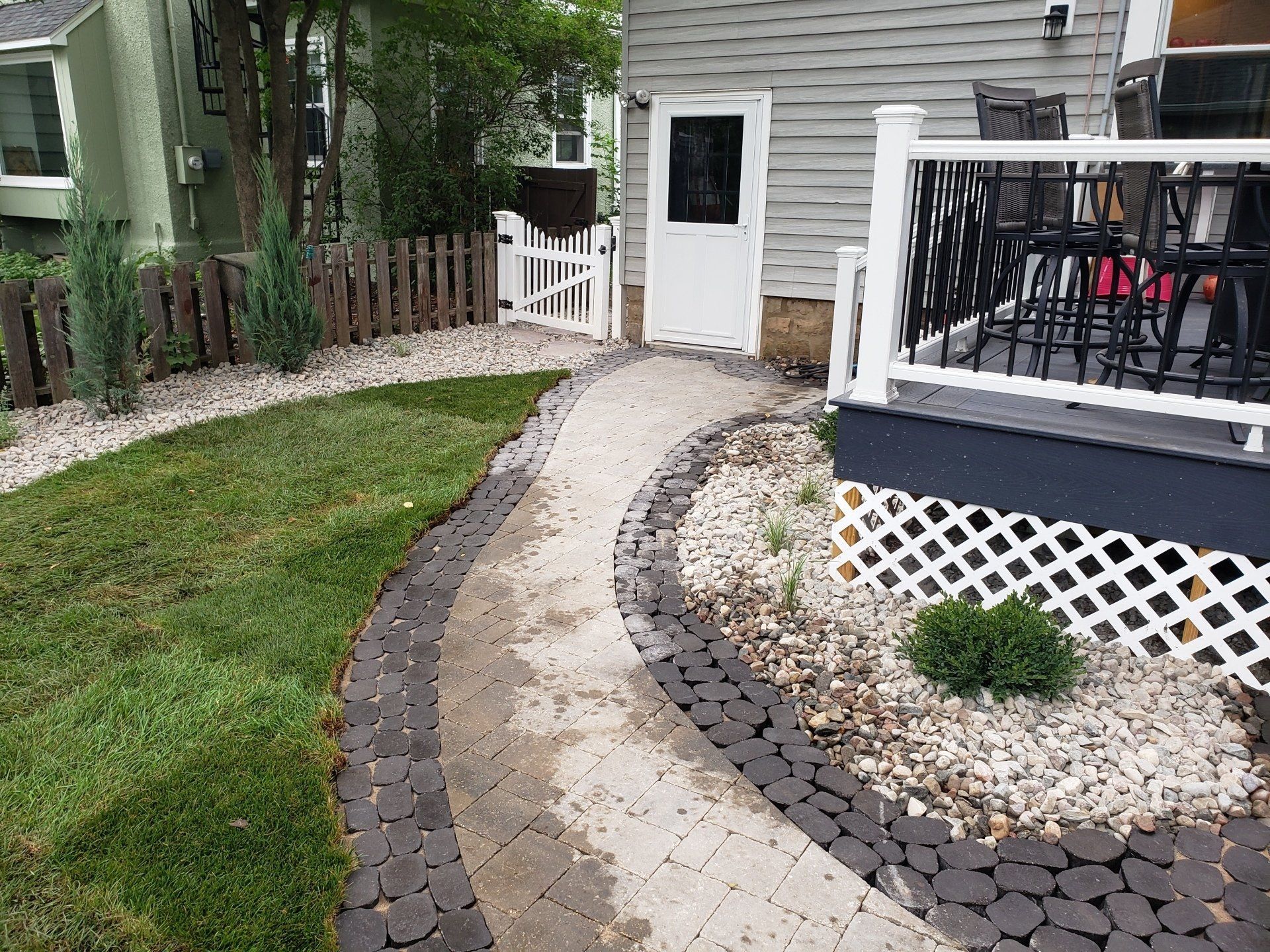 Stone pathway curves through a landscaped yard with green grass, small white gate, and wooden deck.