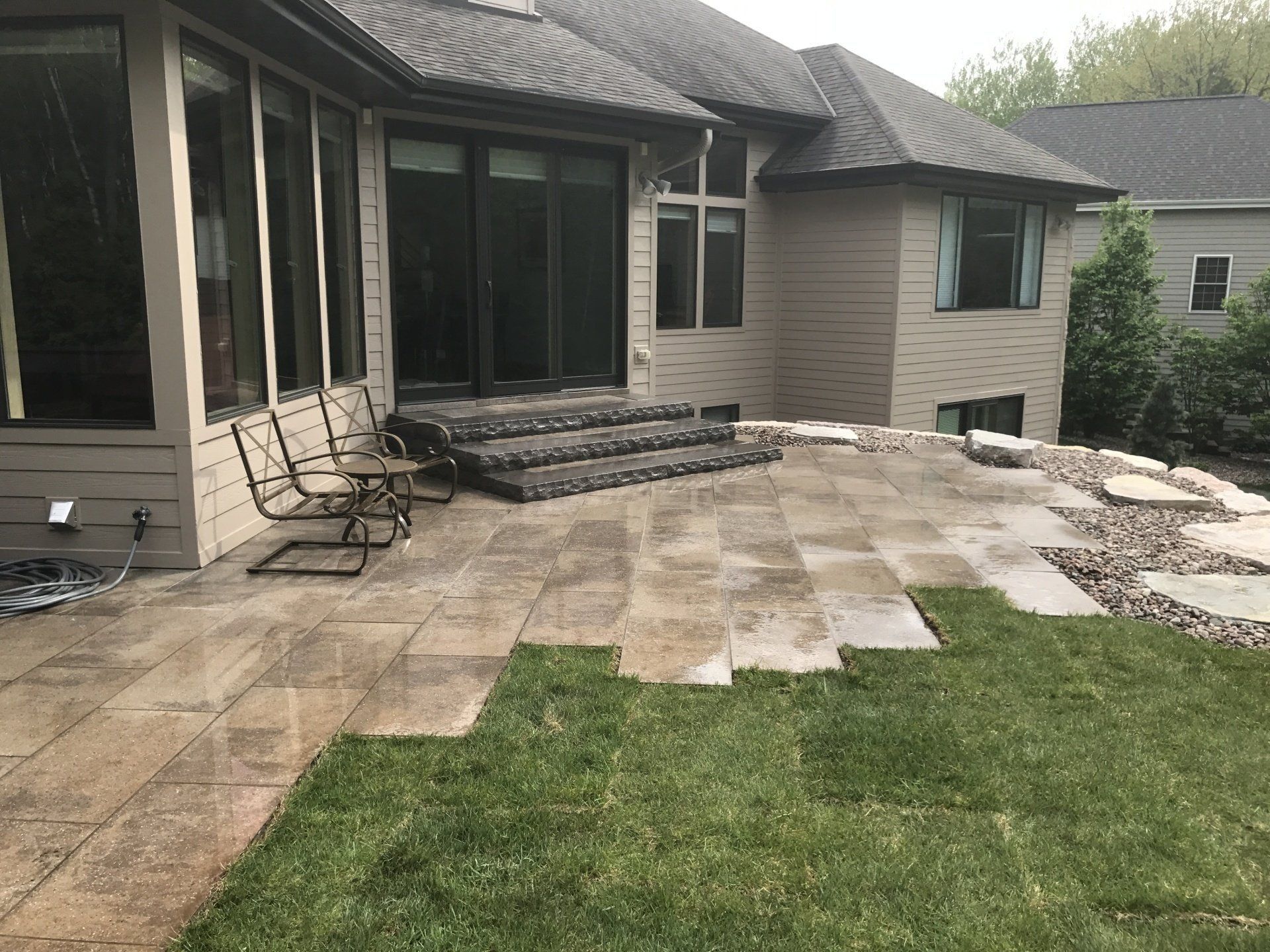 Backyard patio with stone pavers, steps, and lawn, adjacent to a beige house. Two chairs sit on the patio.