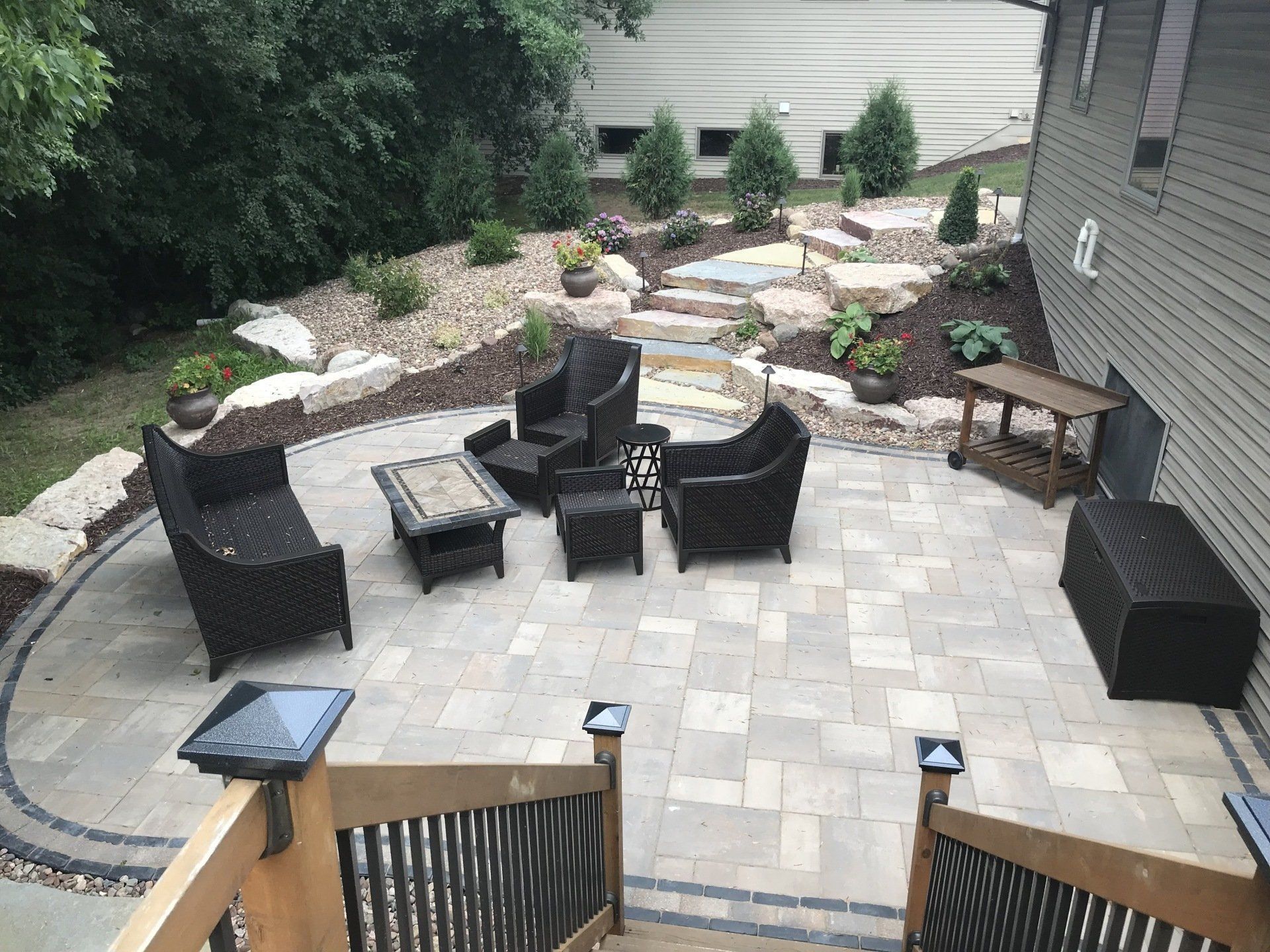 Patio with dark wicker furniture on brick pavers; steps lead down from a wooden deck.