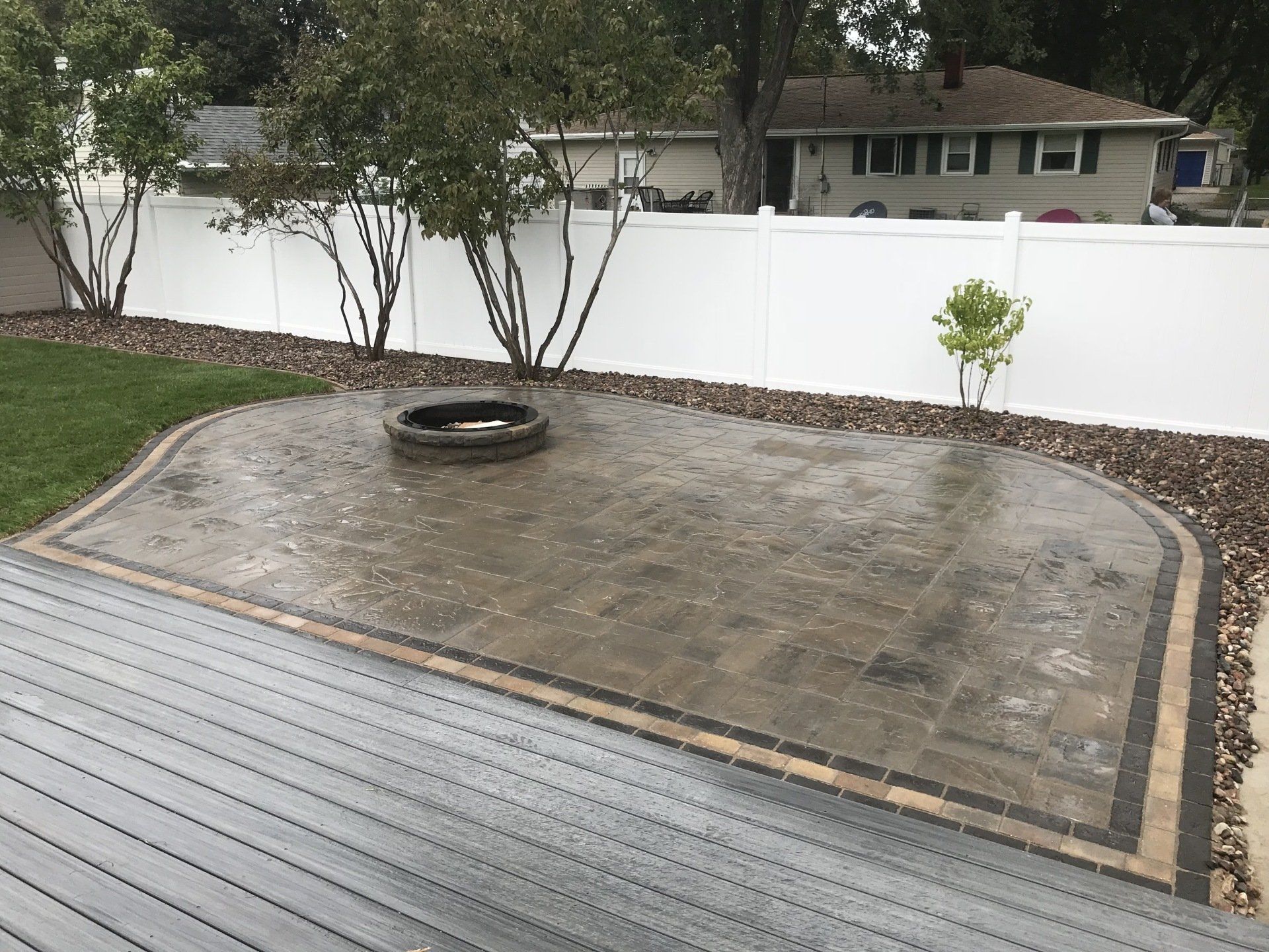 Patio with pavers and fire pit, adjacent to a wooden deck, bordered by a white fence.