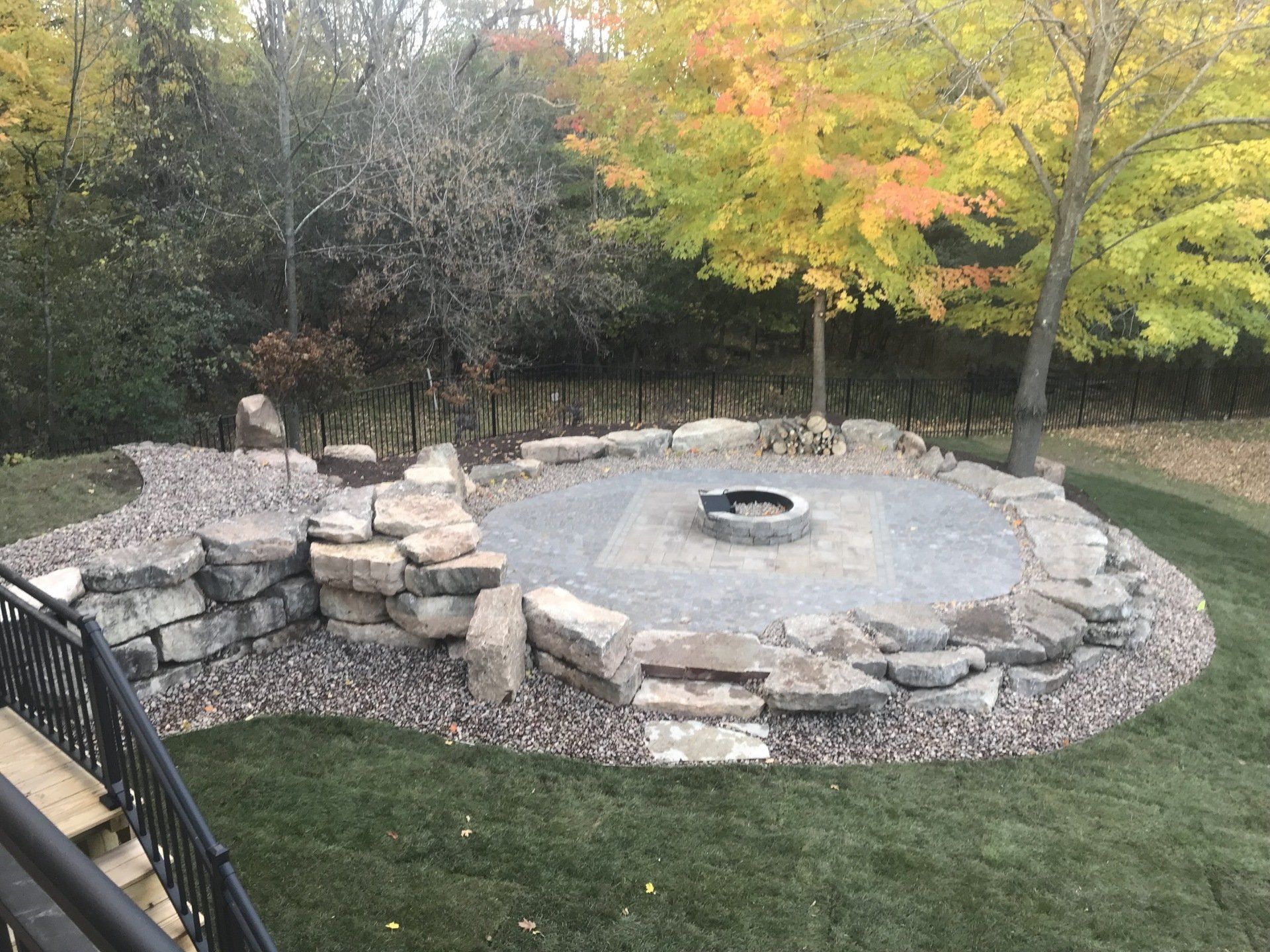 Stone fire pit area on a grassy lawn, surrounded by large rocks and a colorful tree.