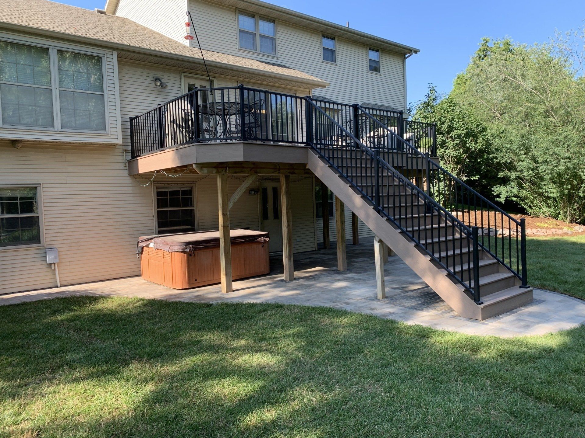 Two-story deck with black railing and stairs, tan siding on a house, hot tub below, and green lawn.