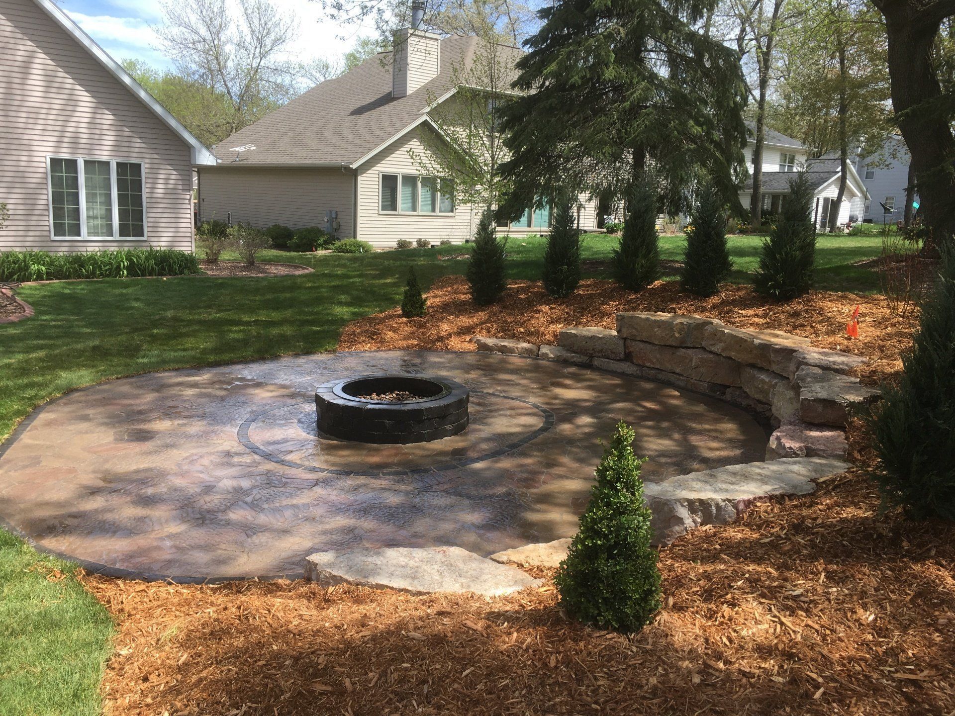 Backyard patio with fire pit, stone wall, and landscaping with mulch and green trees.
