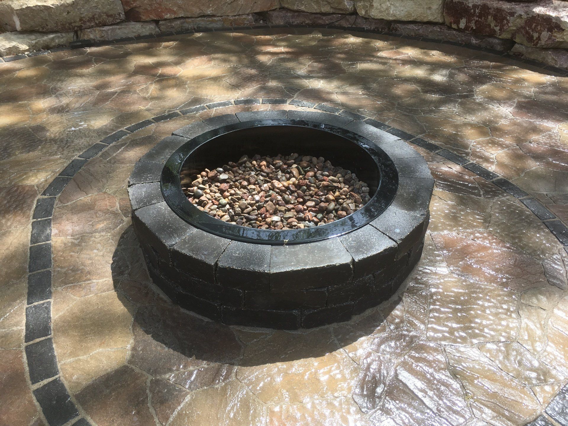 Stone fire pit filled with pebbles, in a paved outdoor area with a decorative border.