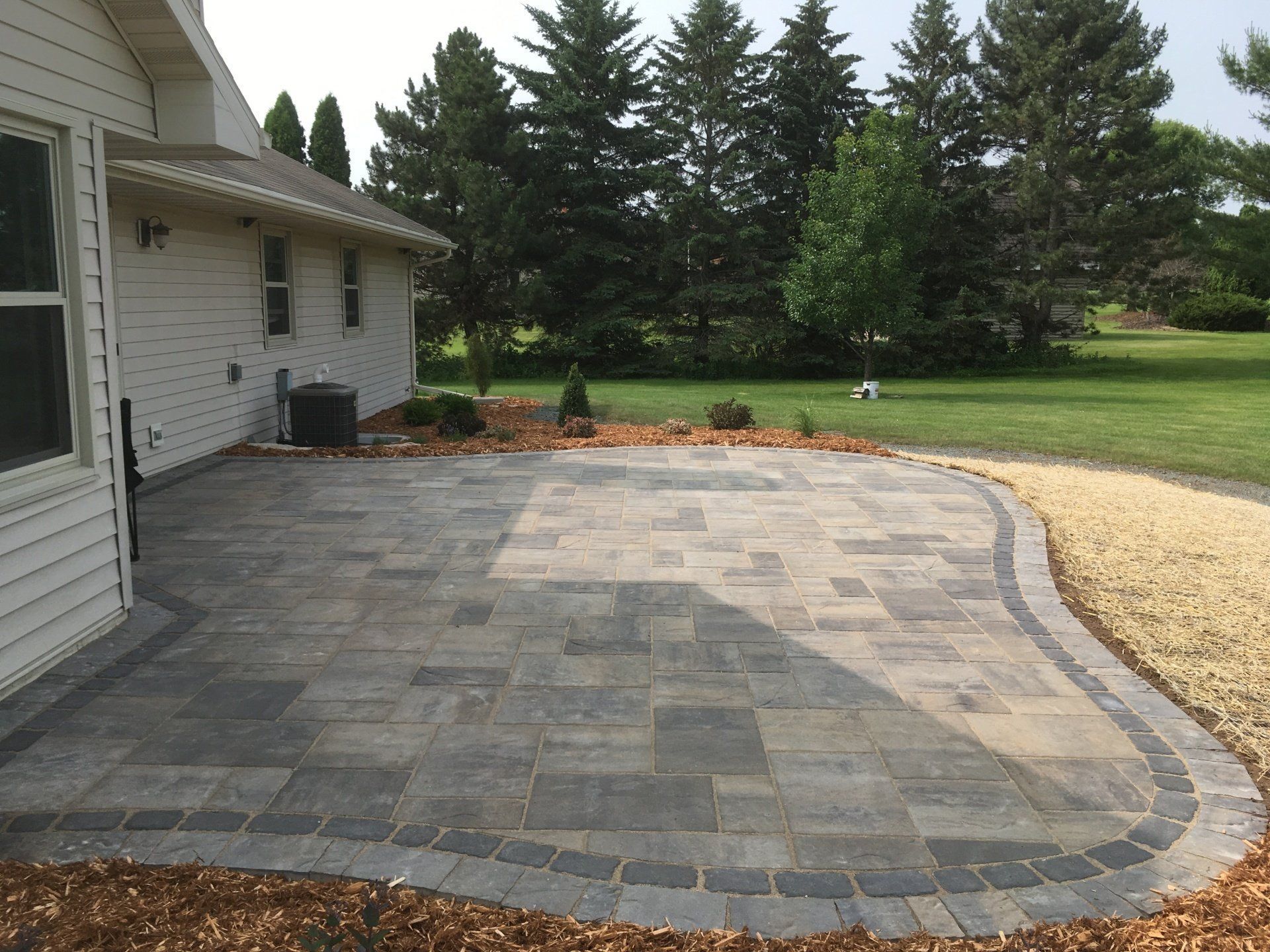 Stone paver patio next to a light-colored house with landscaping and trees in the background.