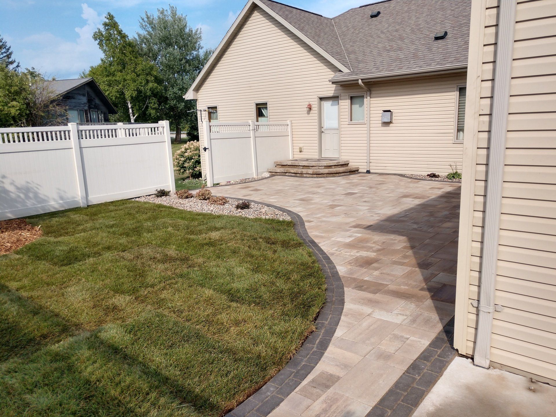 Backyard patio with brick pavers, green grass, and white fence. Beige house in the background.