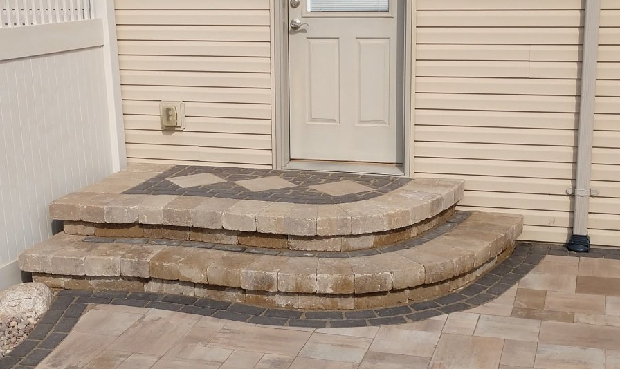 Brick steps leading to a door with a patterned top step, set against a beige siding wall.