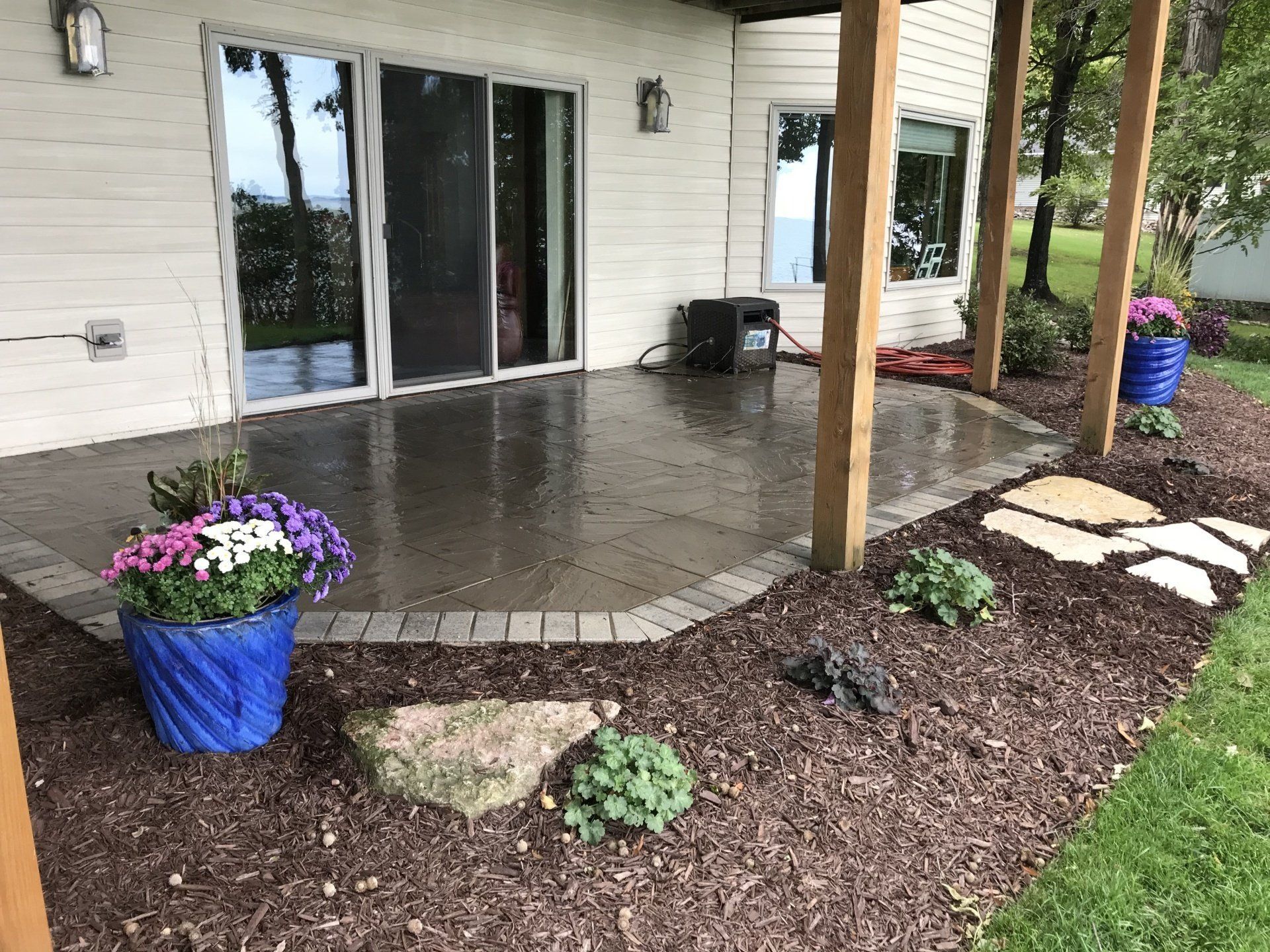 Patio with concrete floor, flower pots, and landscaping. Sliding glass door and wooden support beams visible.