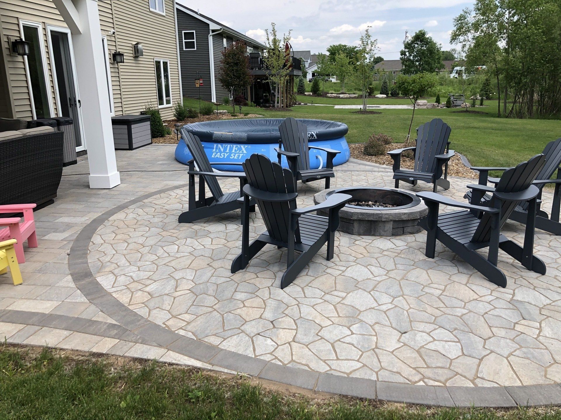 Patio with Adirondack chairs around a fire pit and an above-ground pool in a backyard setting.