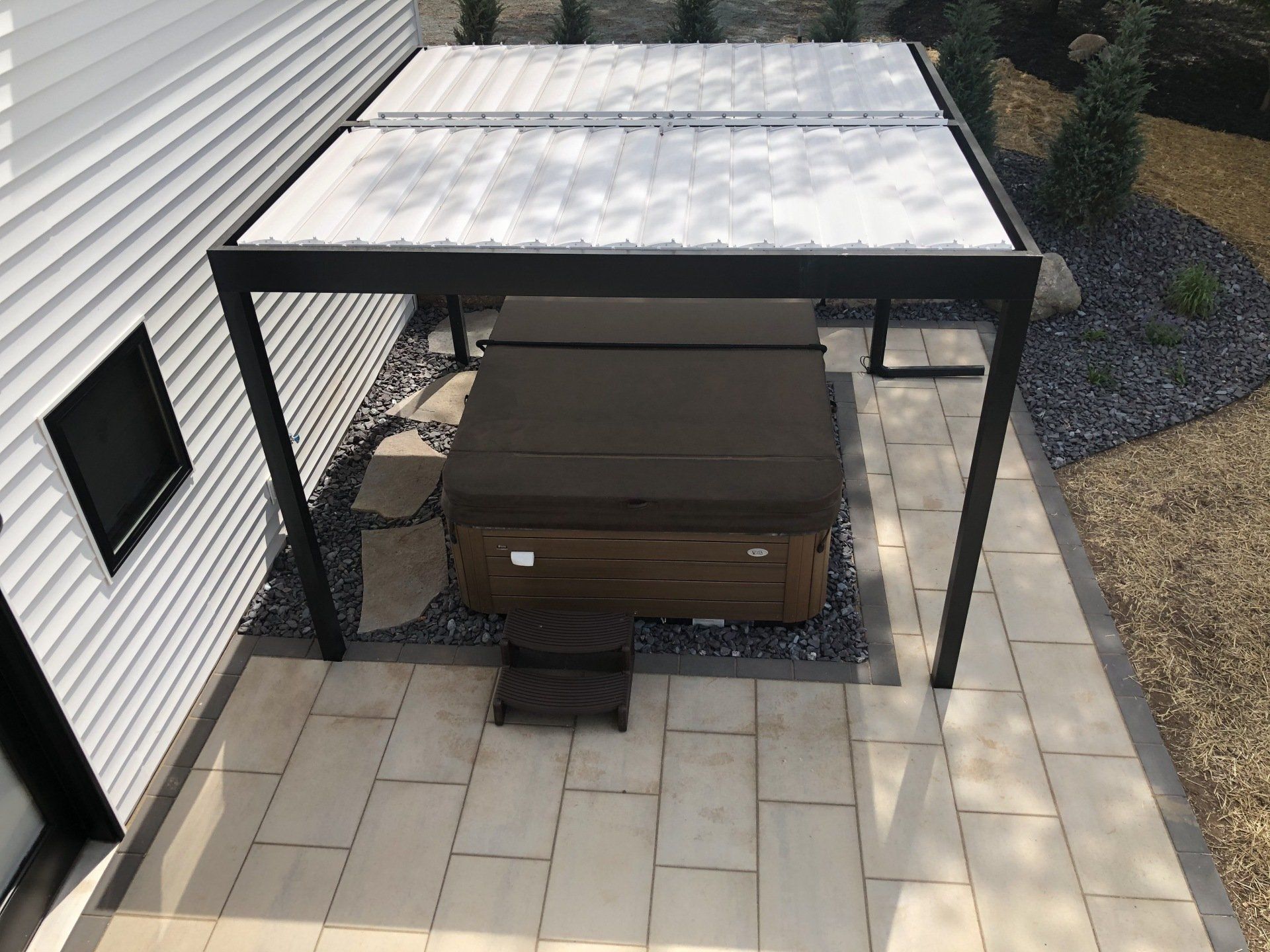 Hot tub under a black pergola on a stone patio next to a white house.