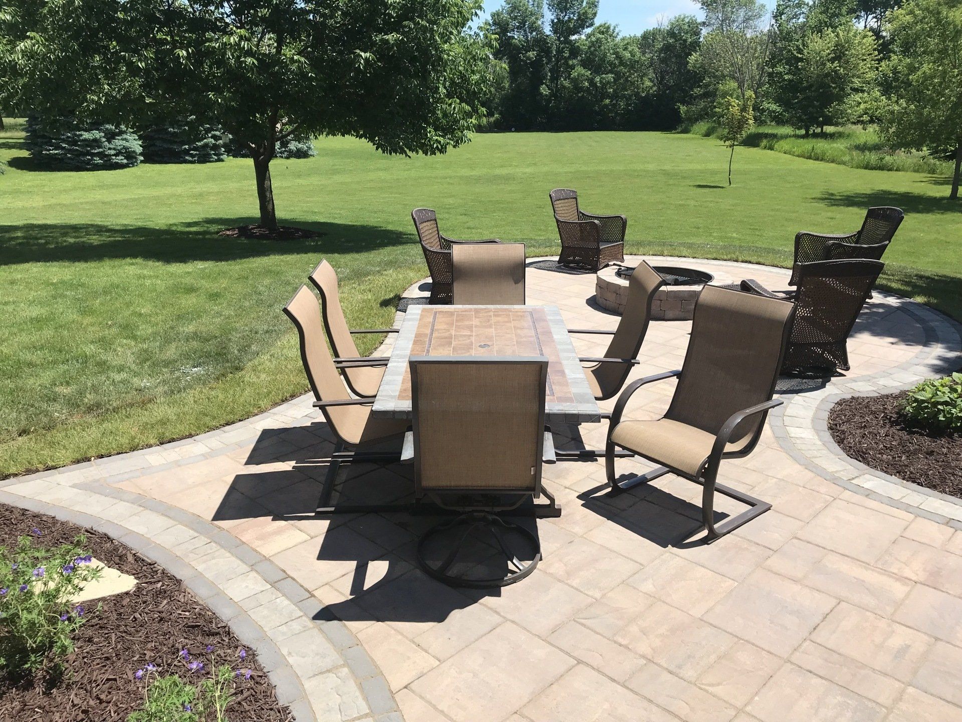 Patio with dining table, chairs, and fire pit on a sunny day in a grassy yard.