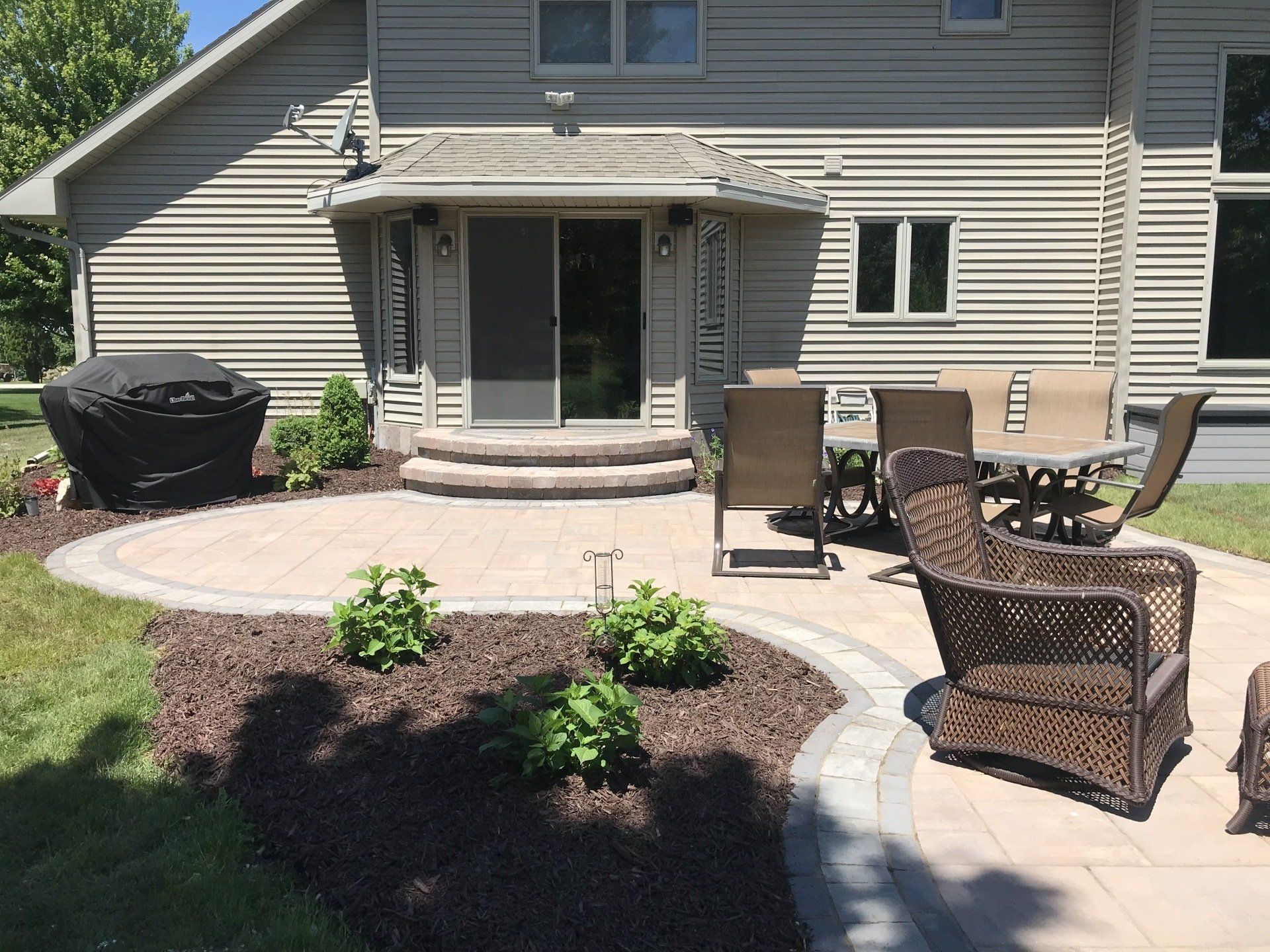 Backyard patio with brick pavers, outdoor dining set, grill, and landscaping. The house is in the background.
