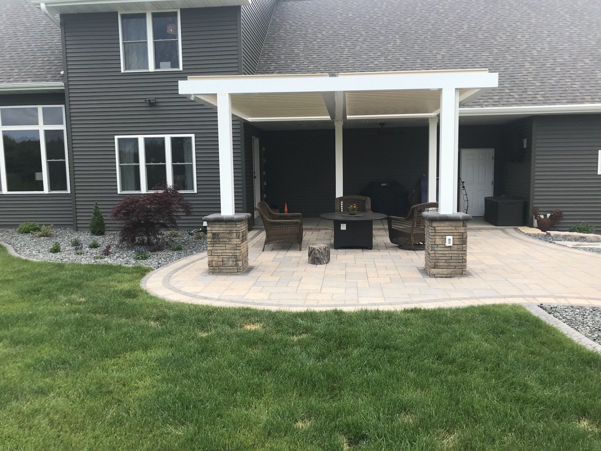 Patio with pergola, seating, fire pit, next to a gray house with windows. Green grass in foreground.