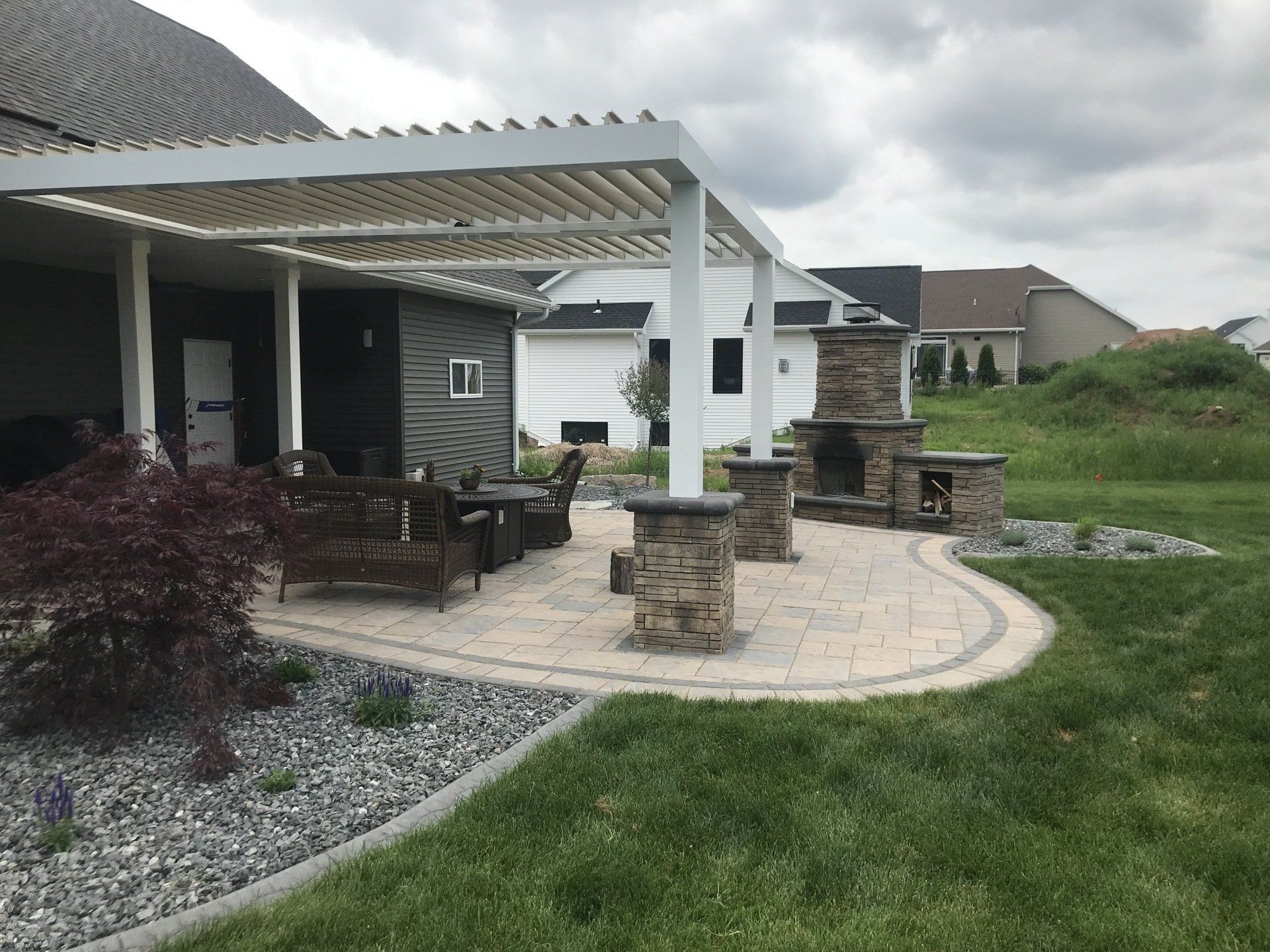 Stone patio with pergola, seating area, fireplace, and surrounding green lawn.