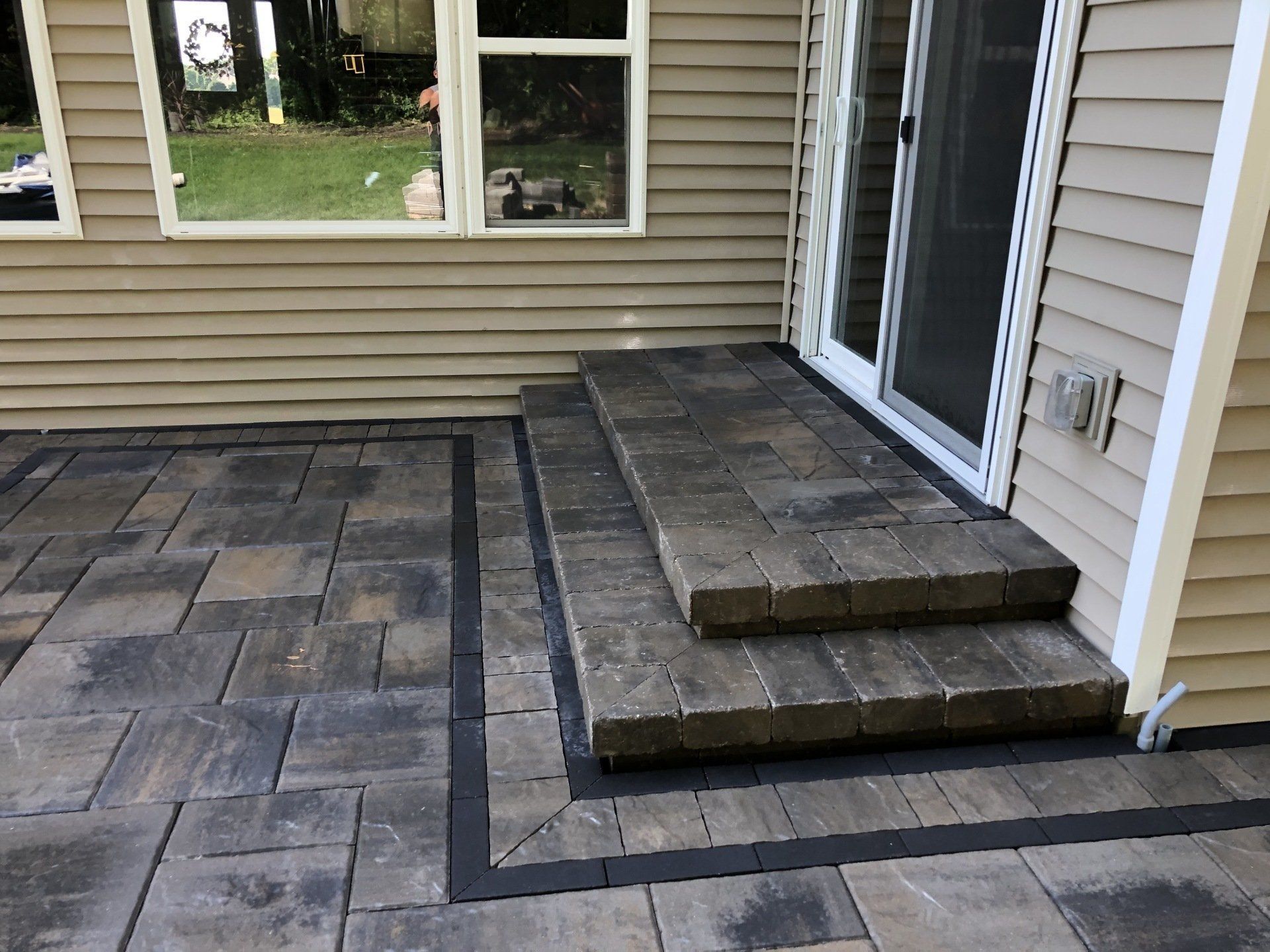 Paver patio with steps leading to a sliding door, bordered in black against a beige house.