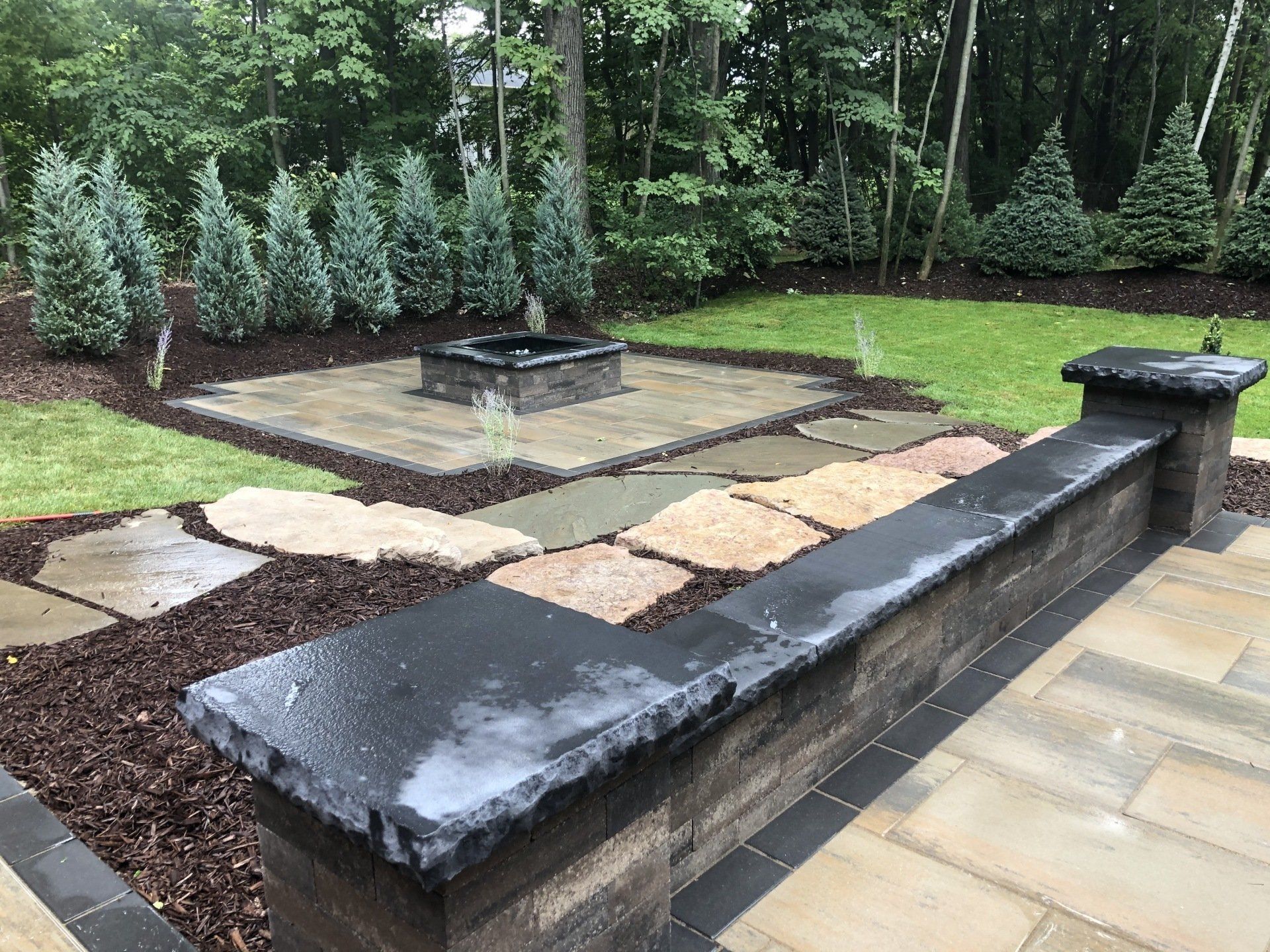 Backyard patio with stone walkway, fire pit, and retaining wall lined with trees.