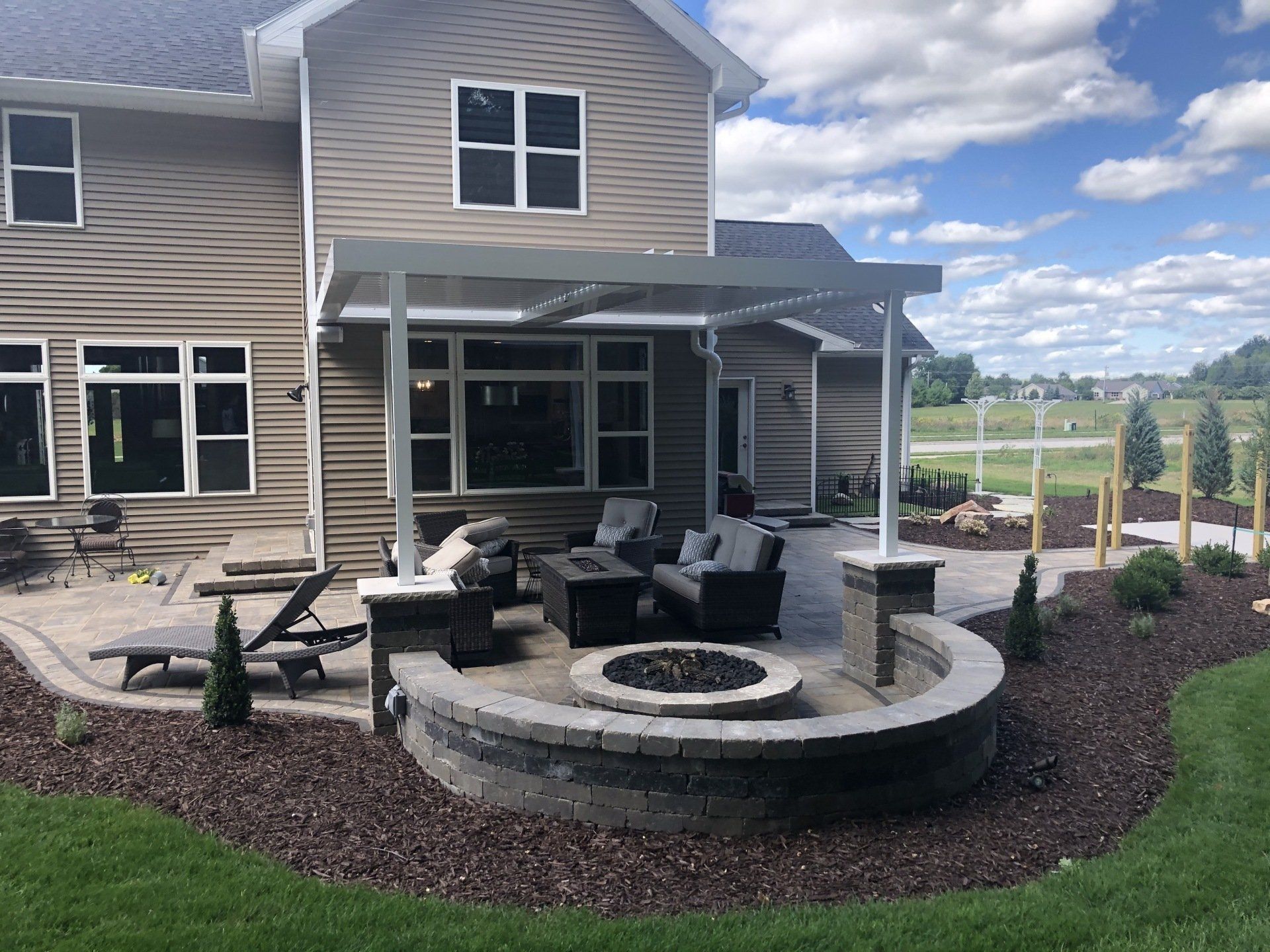 Backyard patio with fire pit and overhead shelter, next to a two-story beige house.