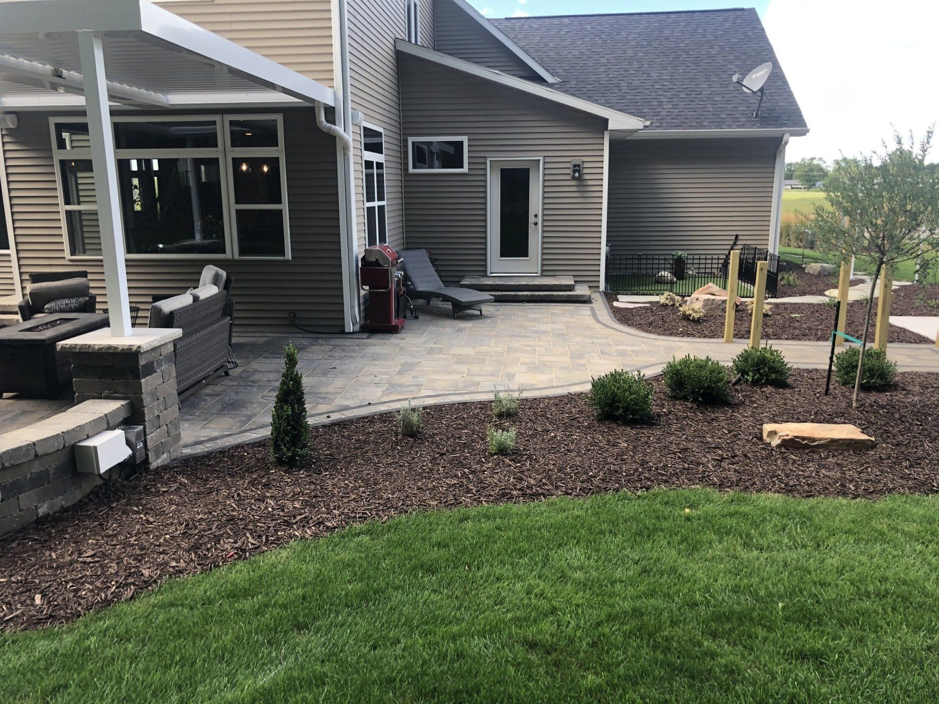 Backyard with patio, brown mulch, green grass, beige siding, and small trees.