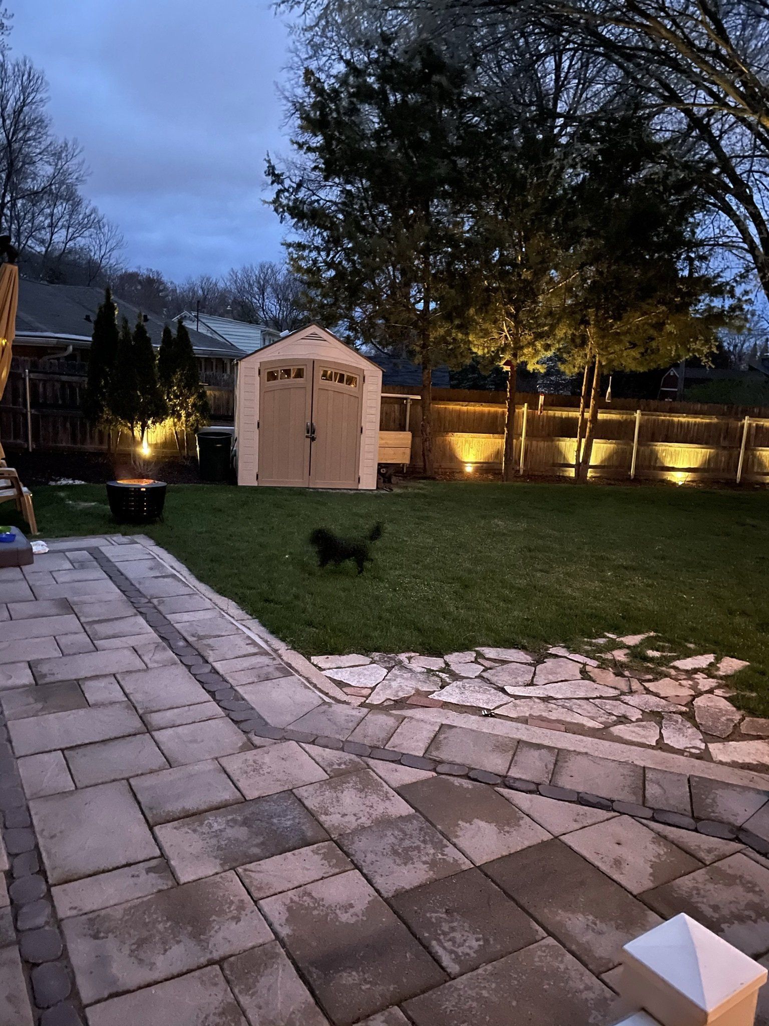 Backyard with a shed, a dog, and stone patio. Lawn and fence are illuminated by lights at dusk.
