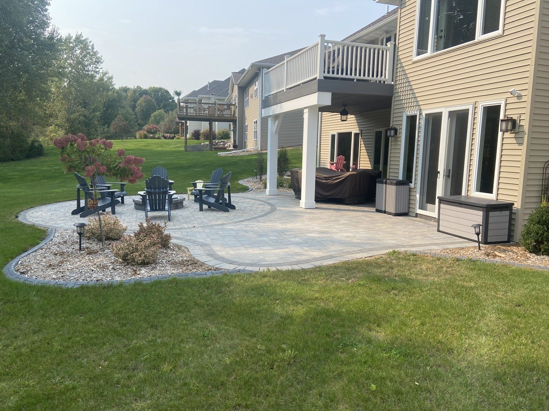 Backyard patio with fire pit, chairs, and house with a deck. Green grass and blue sky.